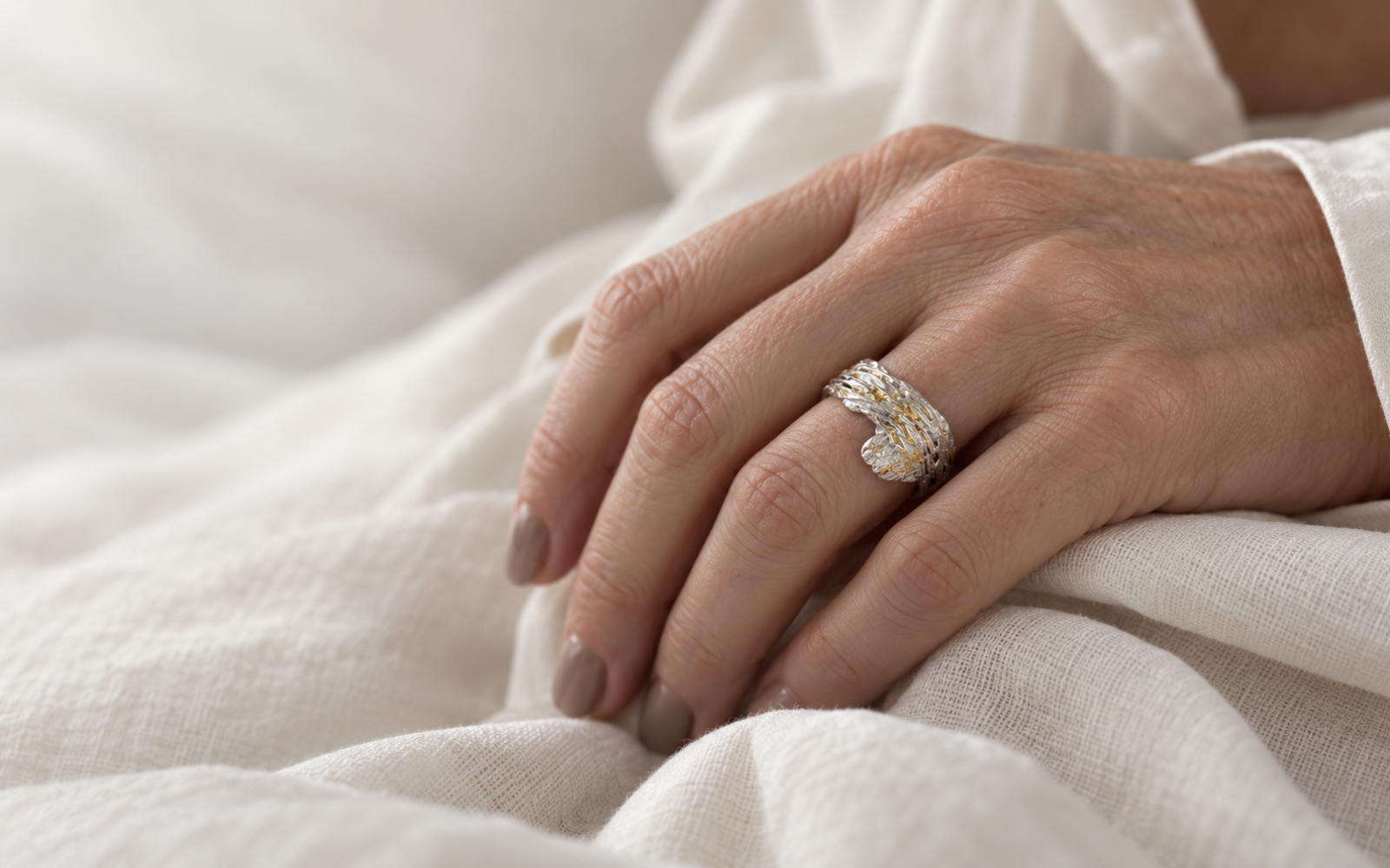 Textured silver ring with gold accents worn on a hand resting on soft off-white fabric.