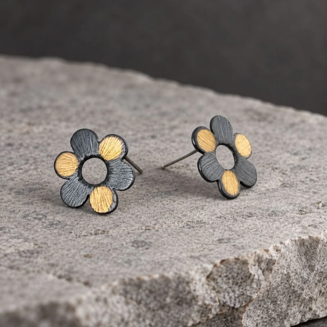Angled view of silver and gold flower stud earrings on gray stone, with posts visible.