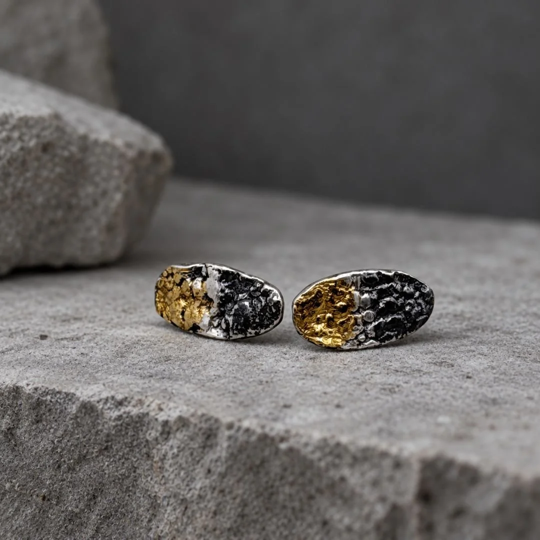 Textured oval stud earrings with contrasting gold and oxidized silver surfaces displayed on gray stone.
