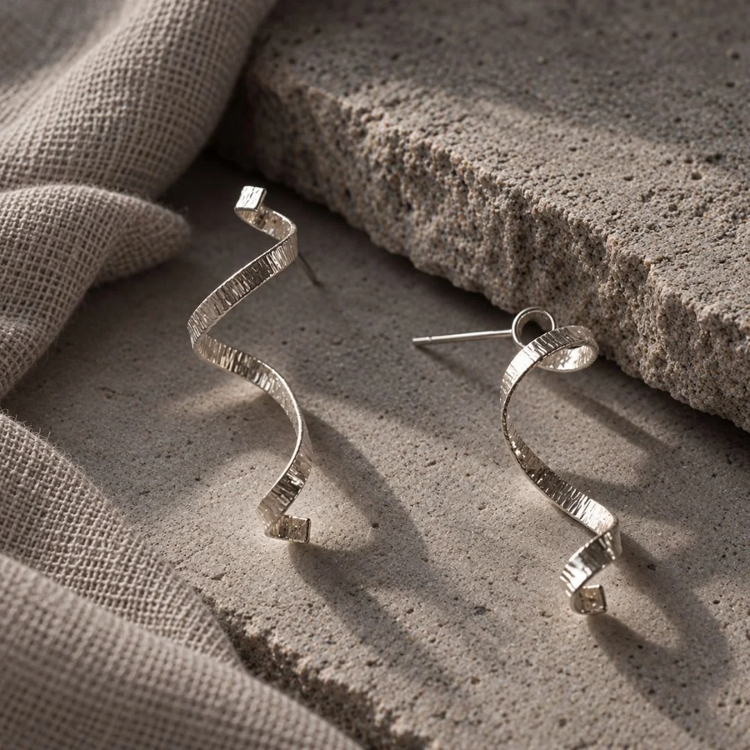 Sterling silver Spiral Ribbon Earrings resting on gray stone beside woven fabric, showing the textured curves and posts.