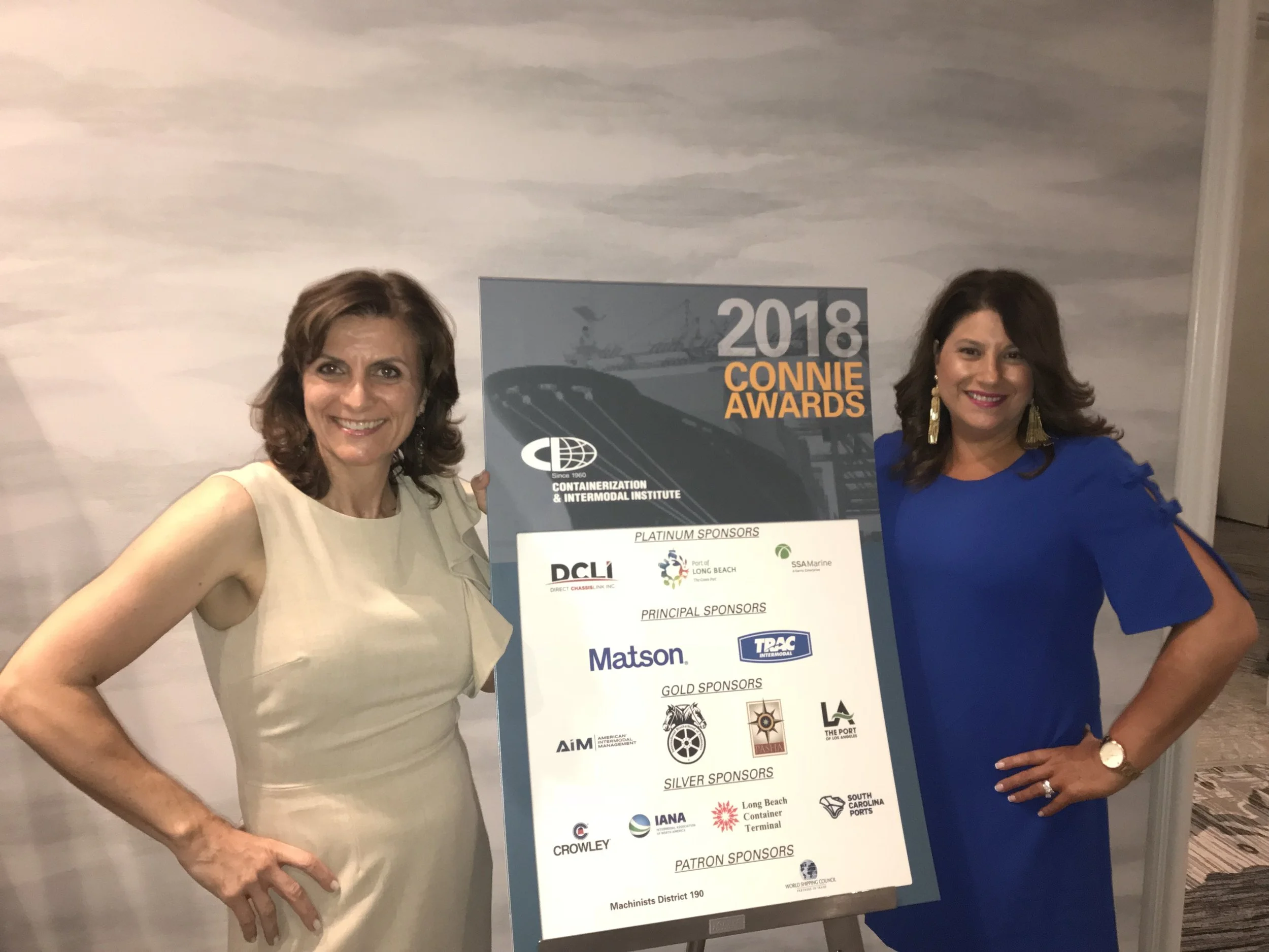 Two women standing next to a sign for the 2018 Connie Awards, highlighting sponsors.