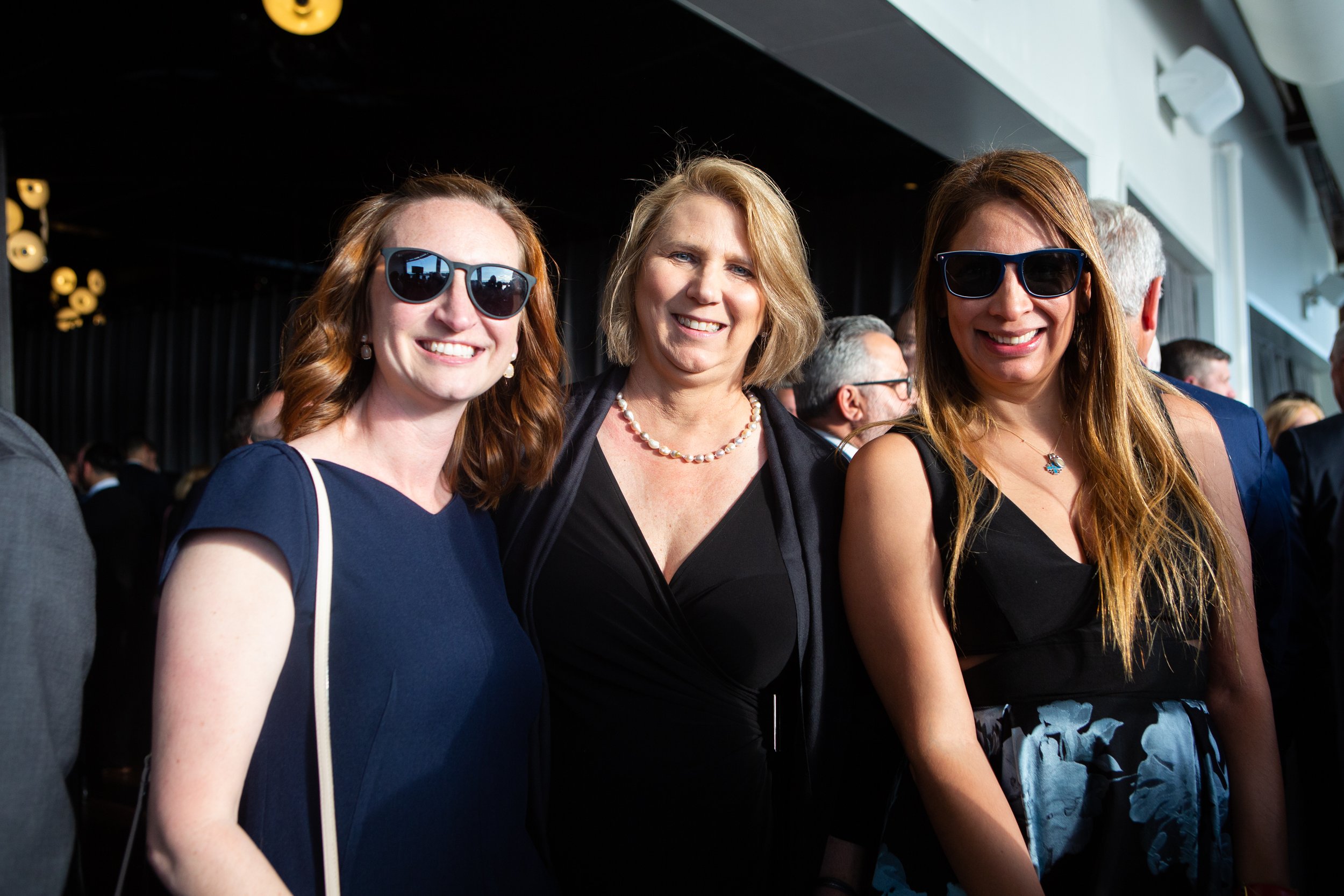 Three women dressed formally, wearing sunglasses, posing together at an indoor event with people in the background.