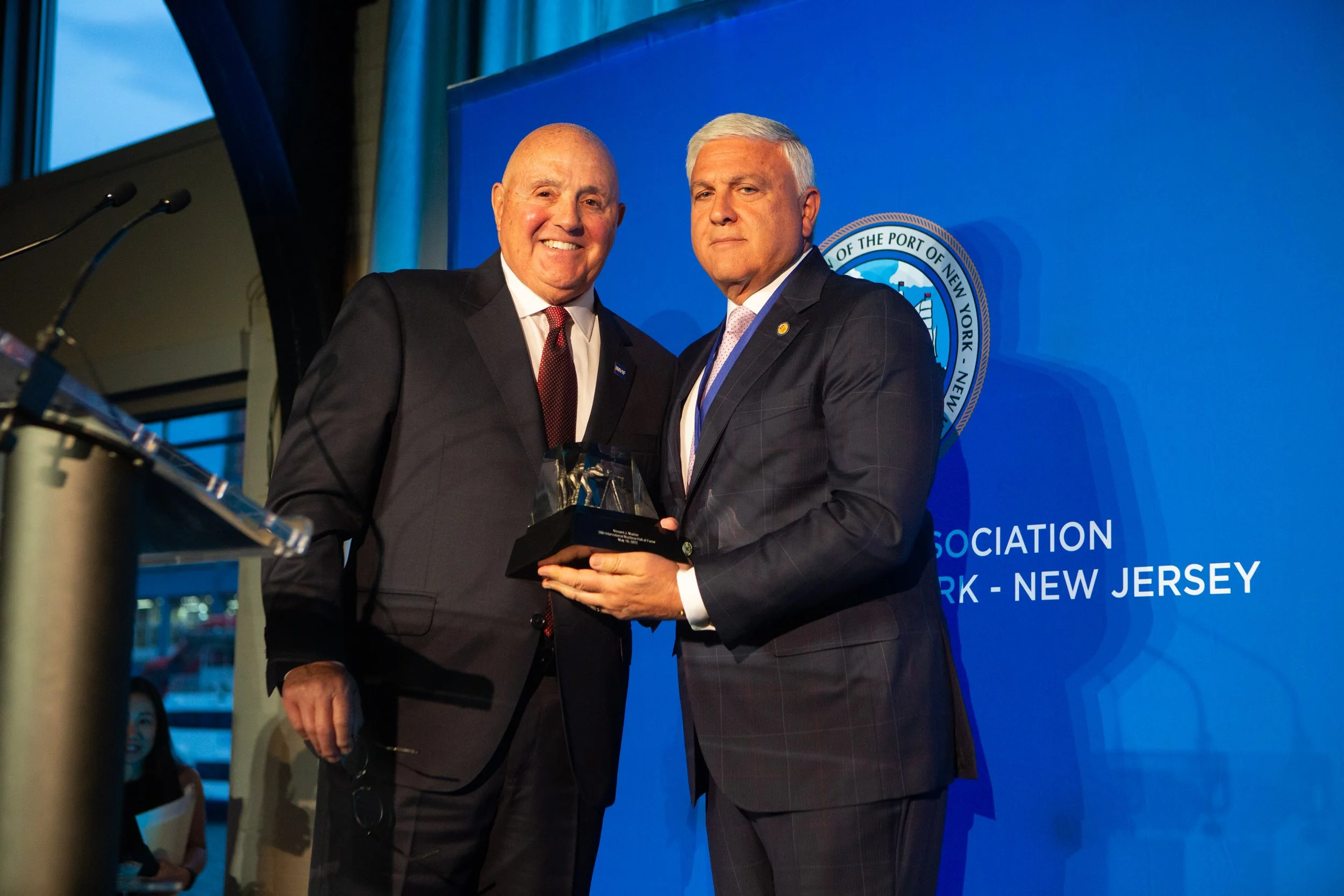 Two men in suits at an event, one holding an award, in front of a blue background with a logo and text about the Port of New York and New Jersey.