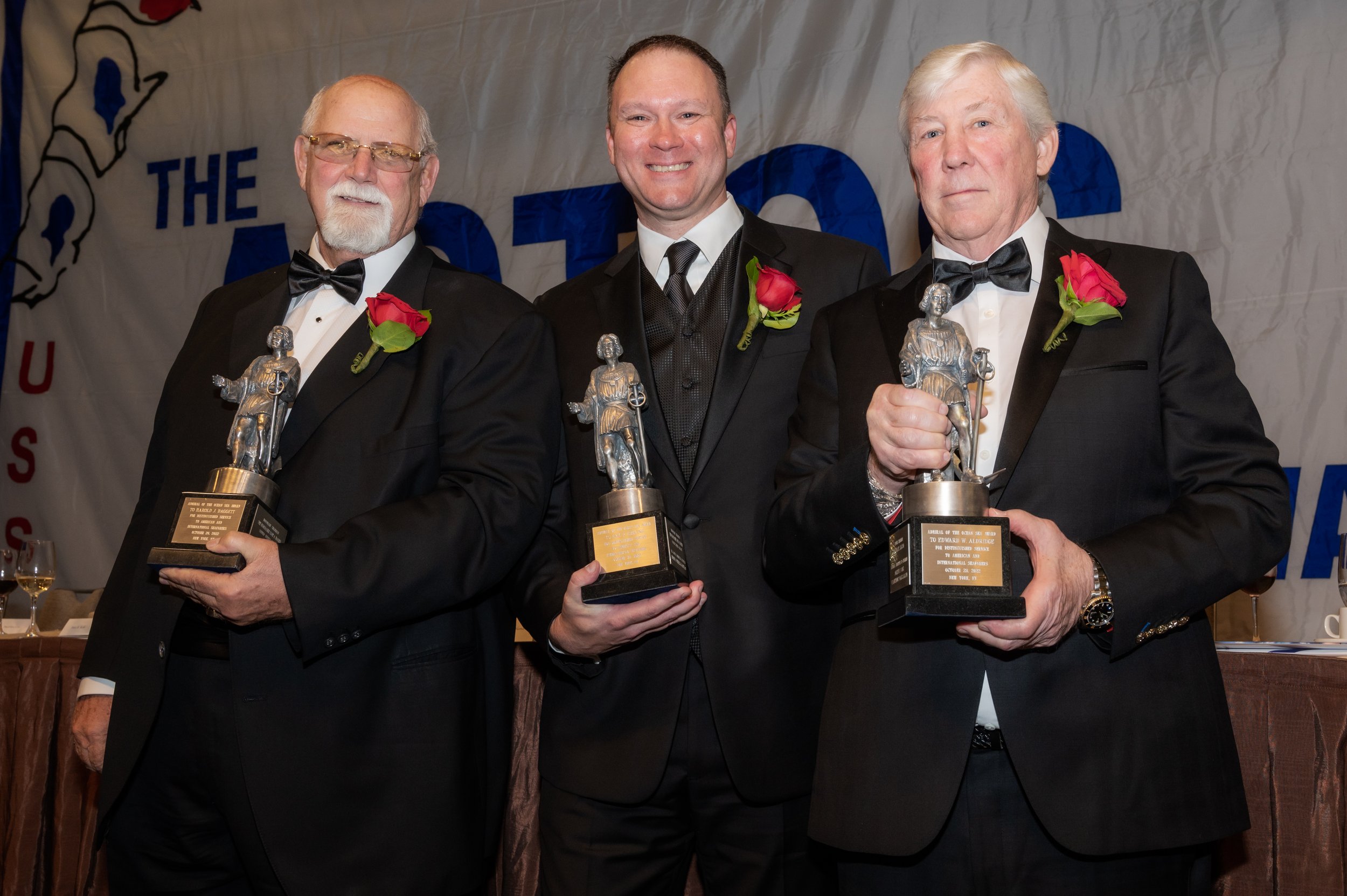 Three men in tuxedos holding awards with red roses on their lapels, standing in front of a banner.