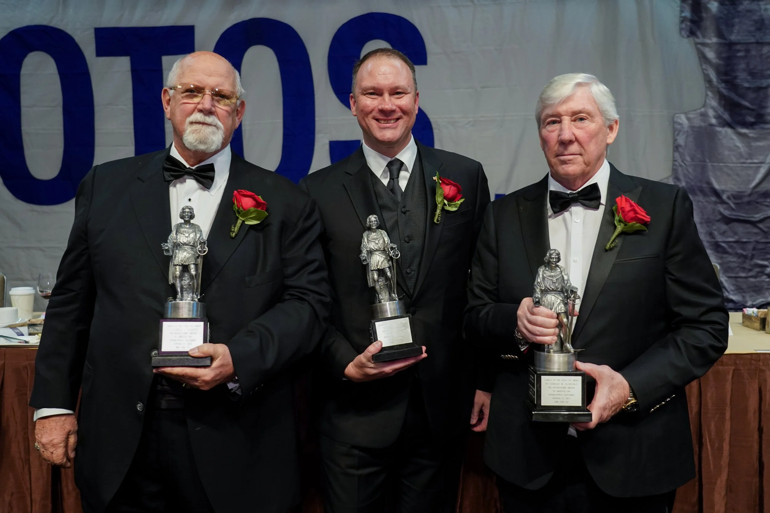 Three men in tuxedos holding trophies with red roses on their lapels.