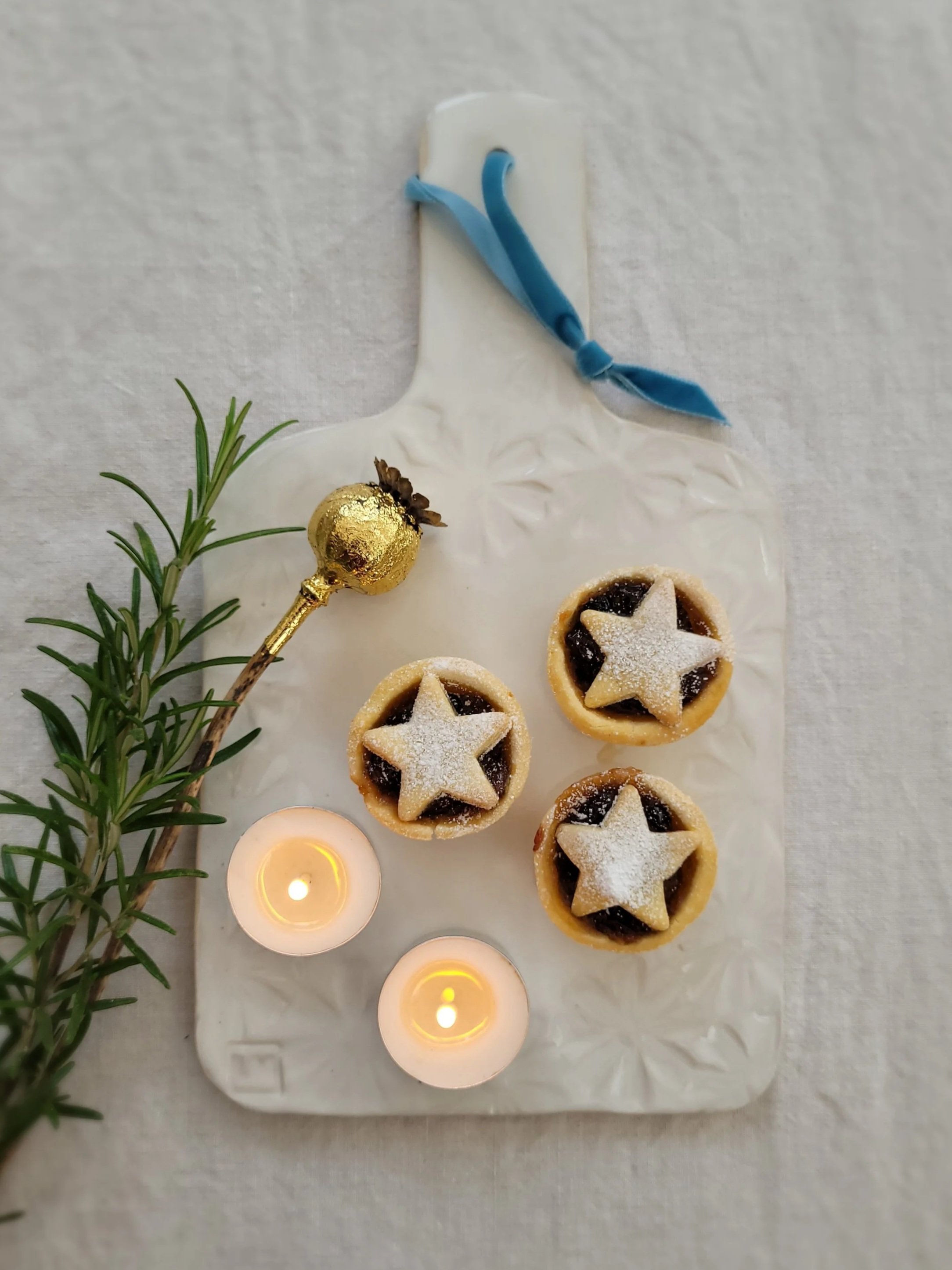 Decorative ceramic tray with three small star-shaped pies dusted with powdered sugar, two lit tealight candles, a sprig of rosemary, and a small gold ornament, all on a light-colored surface.