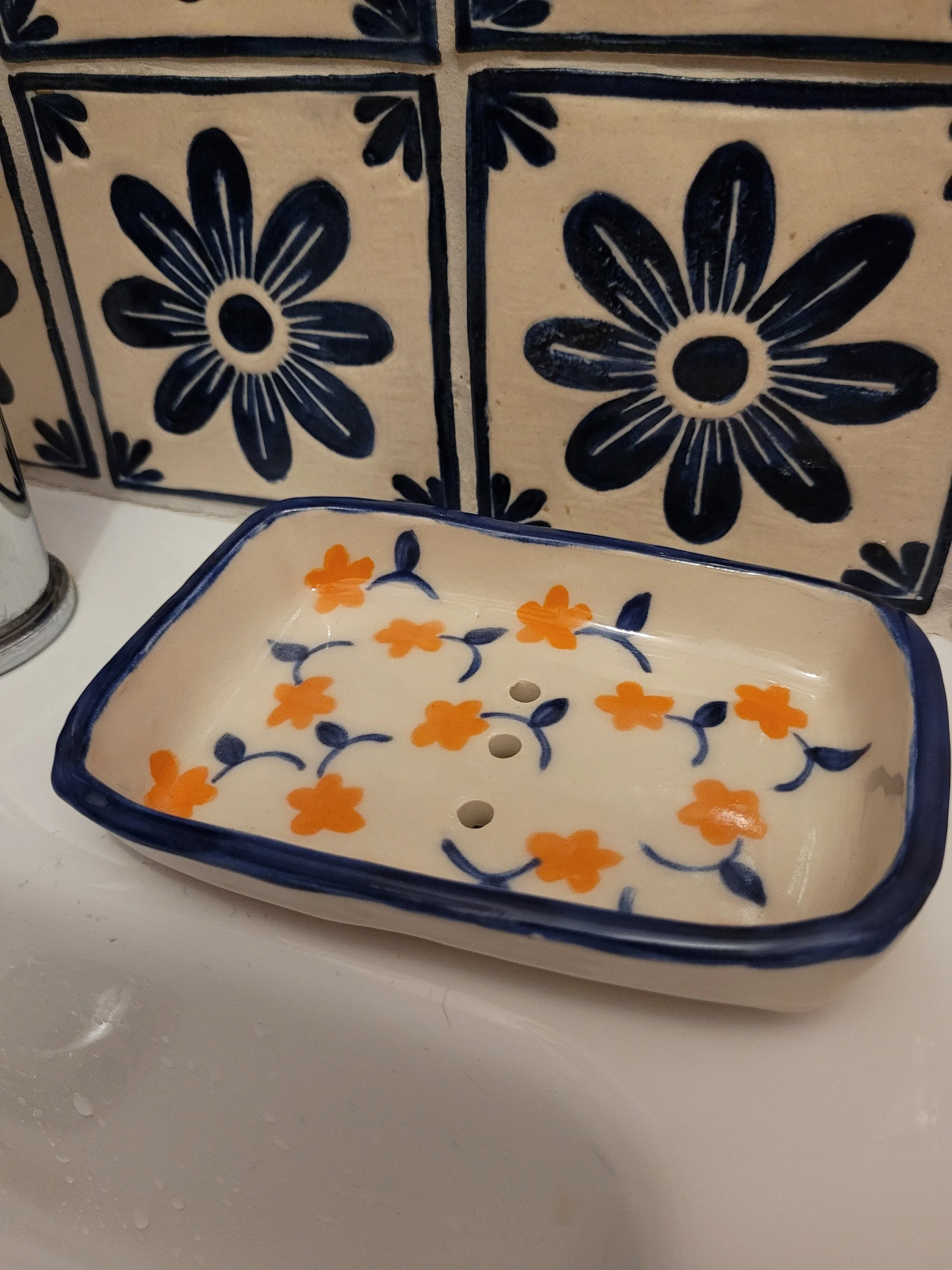 Decorative ceramic soap dish with orange flowers and blue leaves, on a white sink, with a tiled wall featuring large blue flowers in the background.