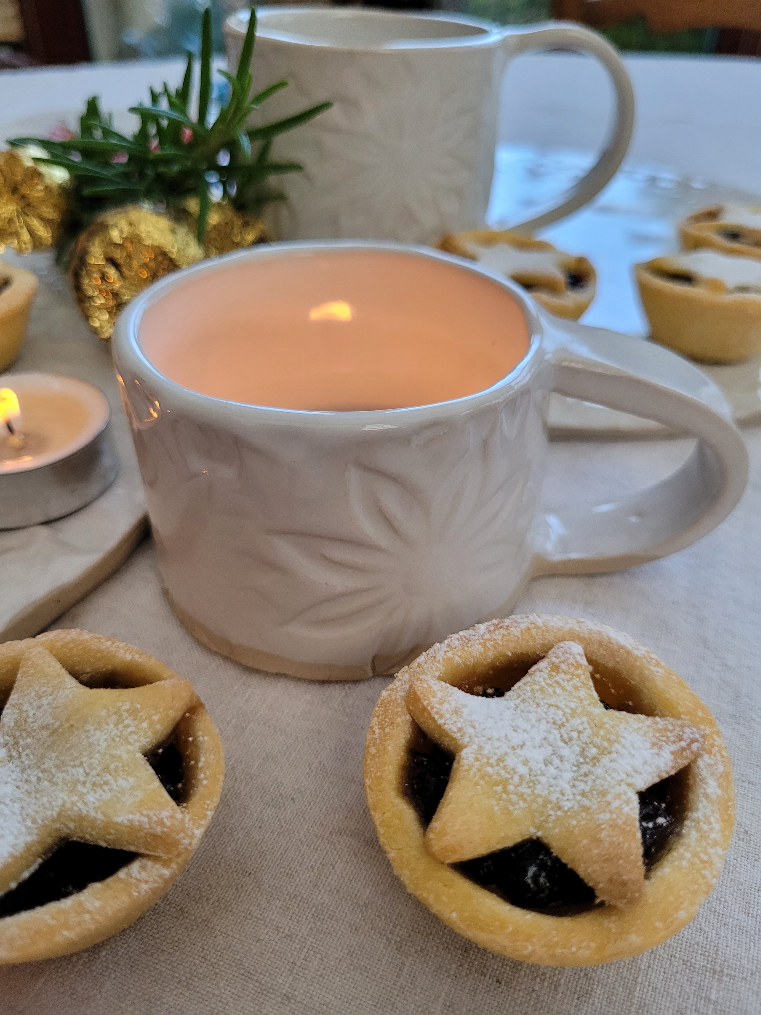 Close-up of a holiday table with a lit candle in a white mug, star-shaped fruit pies with powdered sugar, and festive decorations in the background.