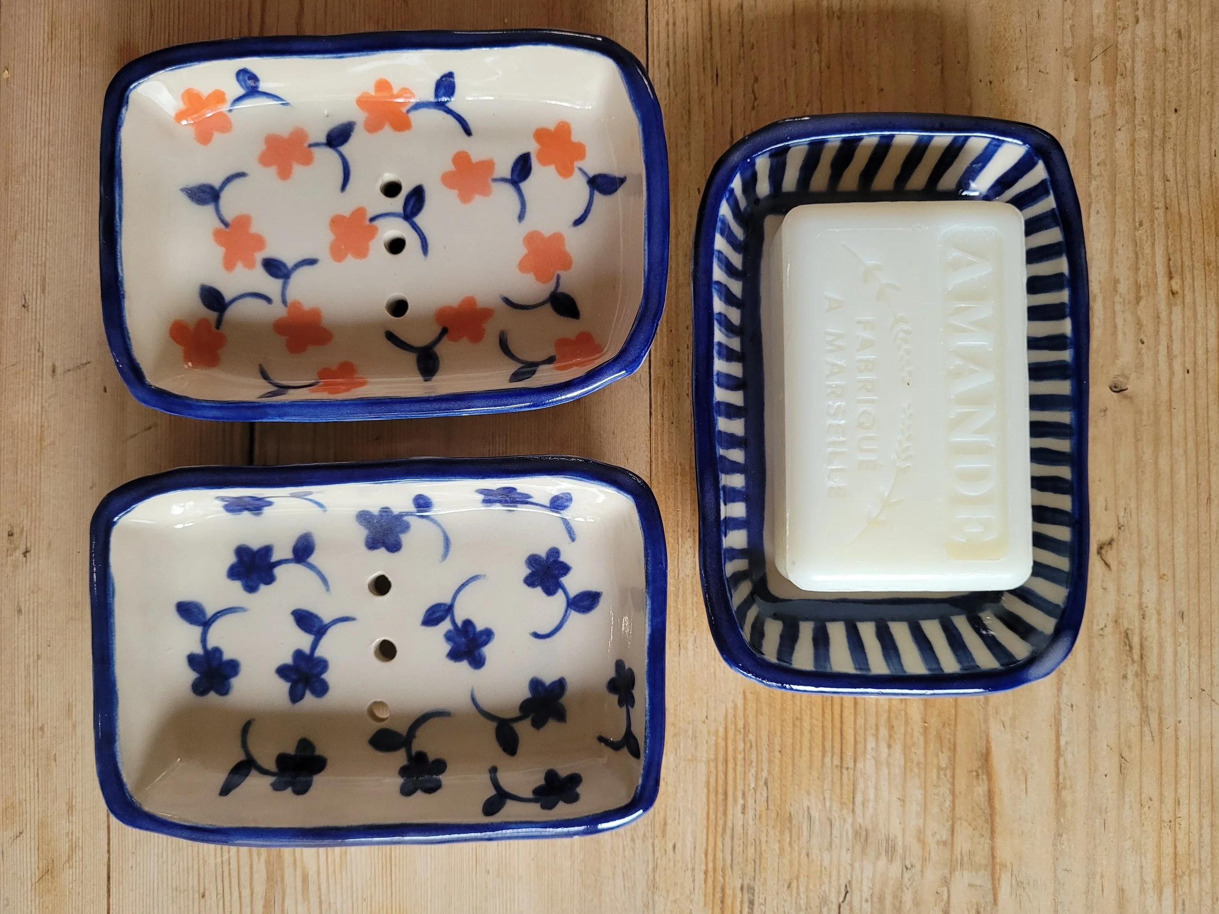Two square ceramic dishes with floral patterns and holes at the bottom, and one rectangular ceramic dish with blue and white stripes holding a block of ivory-colored soap.