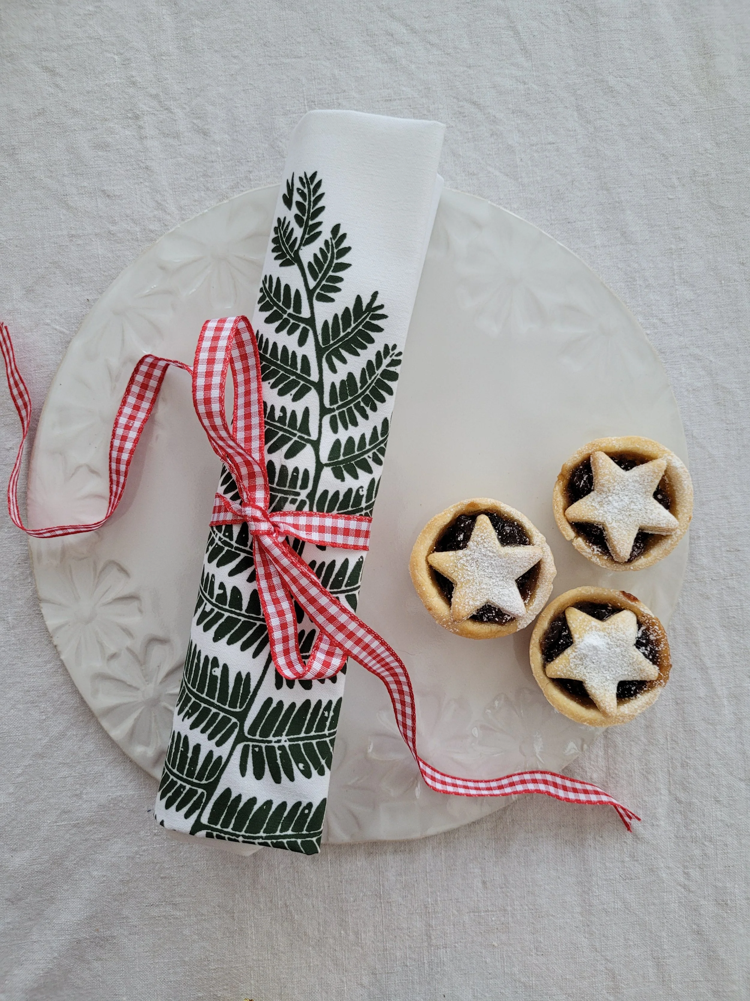 A decorative white plate with three small berry tarts topped with star-shaped pastry cutouts. A paper napkin with a green leaf pattern, tied with a red gingham ribbon, is also on the plate.