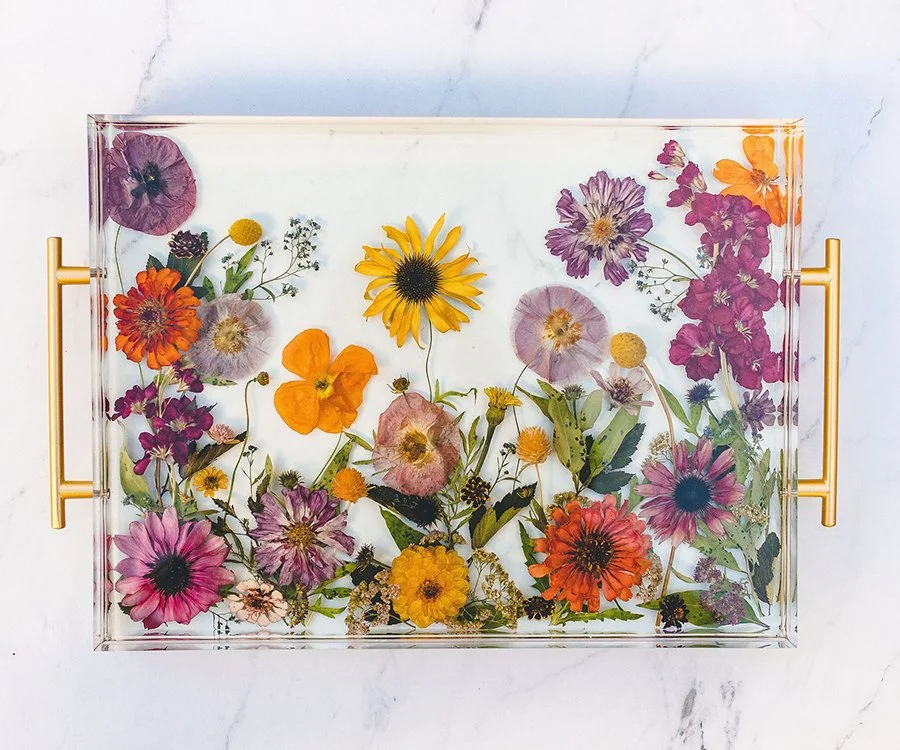 A glass tray filled with colorful pressed flowers, including zinnias, poppies, daisies, and other wildflowers, with gold handles on a white surface.