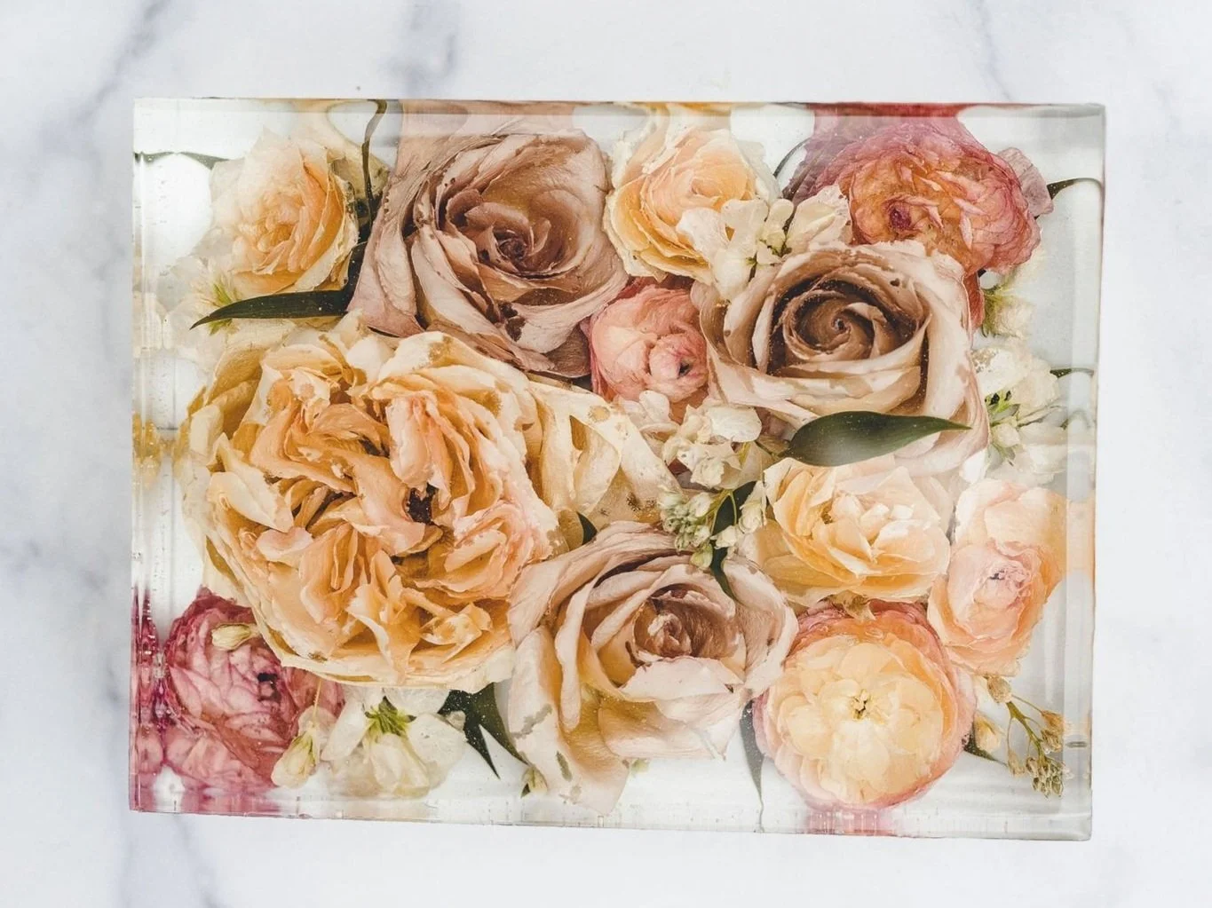 Arranged in a glass container with water, various peach, cream, and pink roses, along with small white flowers and green leaves.