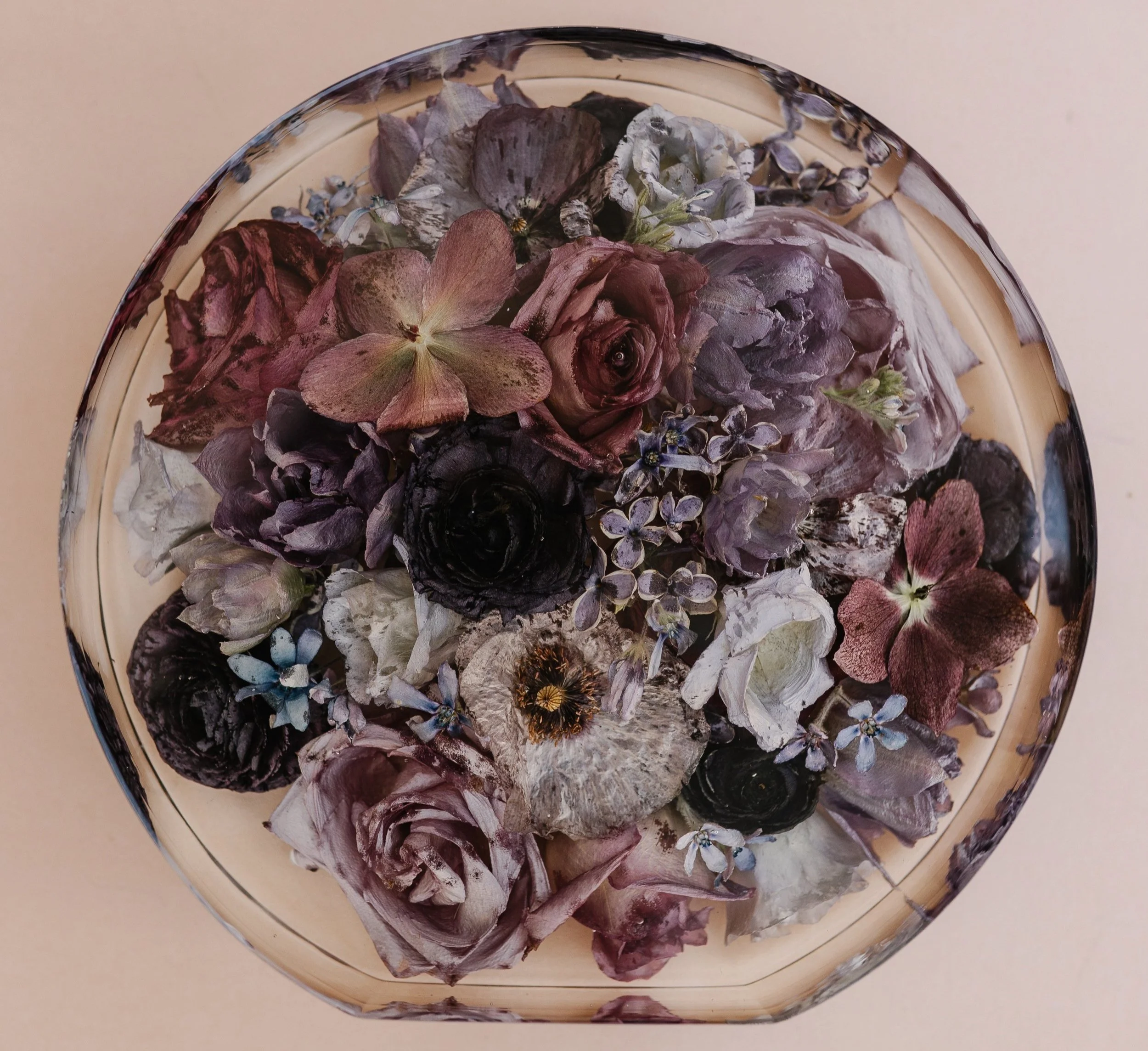 A glass bowl filled with dried purple, white, and pink flowers, including roses and hydrangeas, on a light pink background.