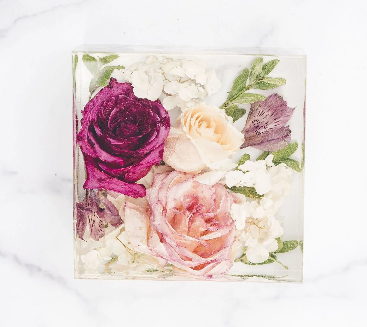 Square glass container with dried pink, purple, cream, and white roses, and white hydrangea flowers, with green leaves against a white background.