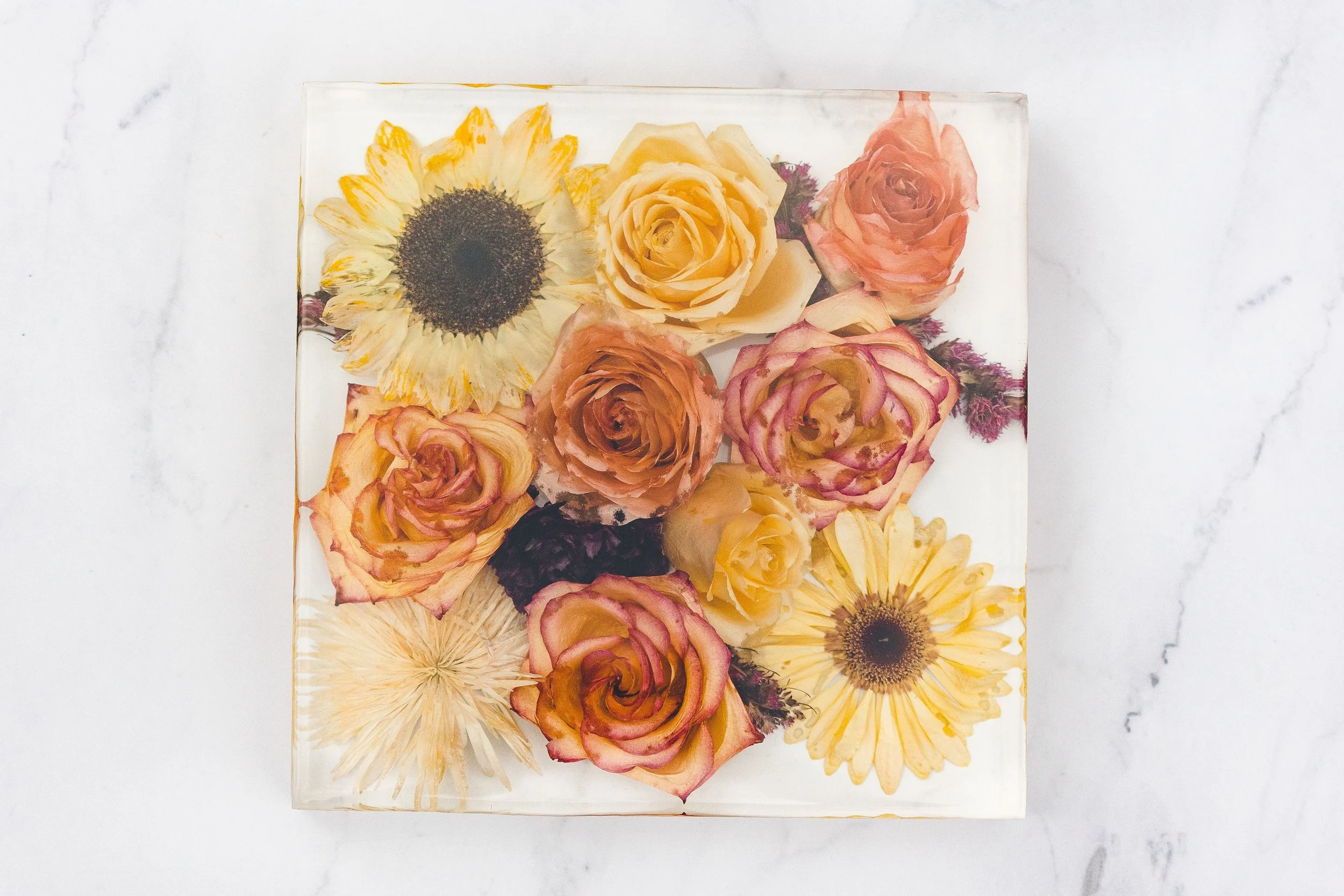 Top view of a square glass box filled with yellow and pink roses, sunflowers, and other flowers on a white marble surface.