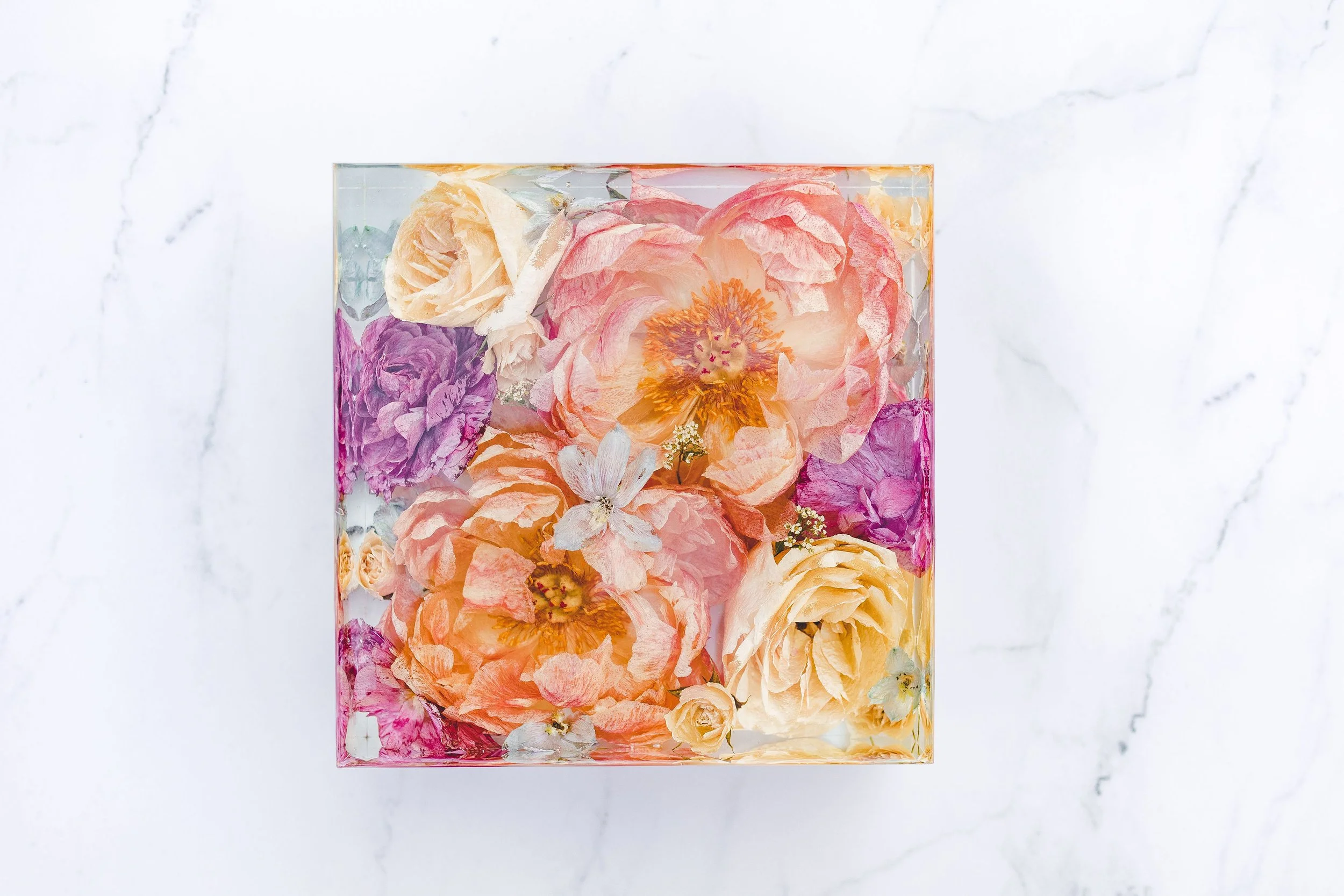 A square glass container filled with various colorful dried flowers against a white marble background.