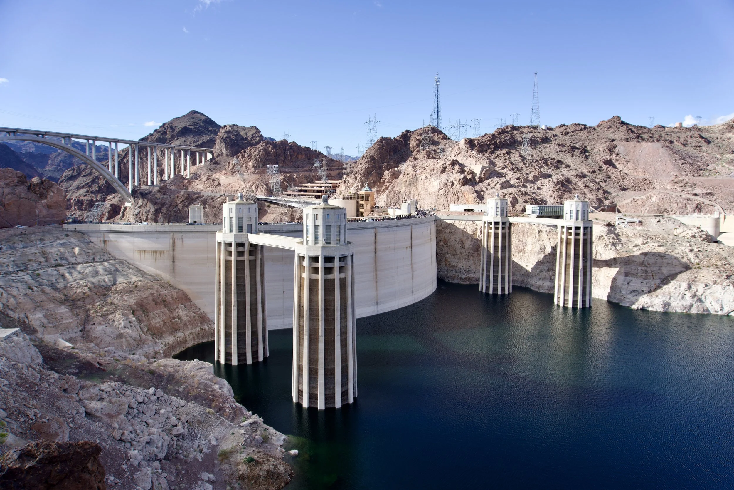 Image of the Hoover Dam with its large concrete arch-gravity dam spanning the Colorado River in a rocky desert landscape, with transmission towers and a bridge in the background.