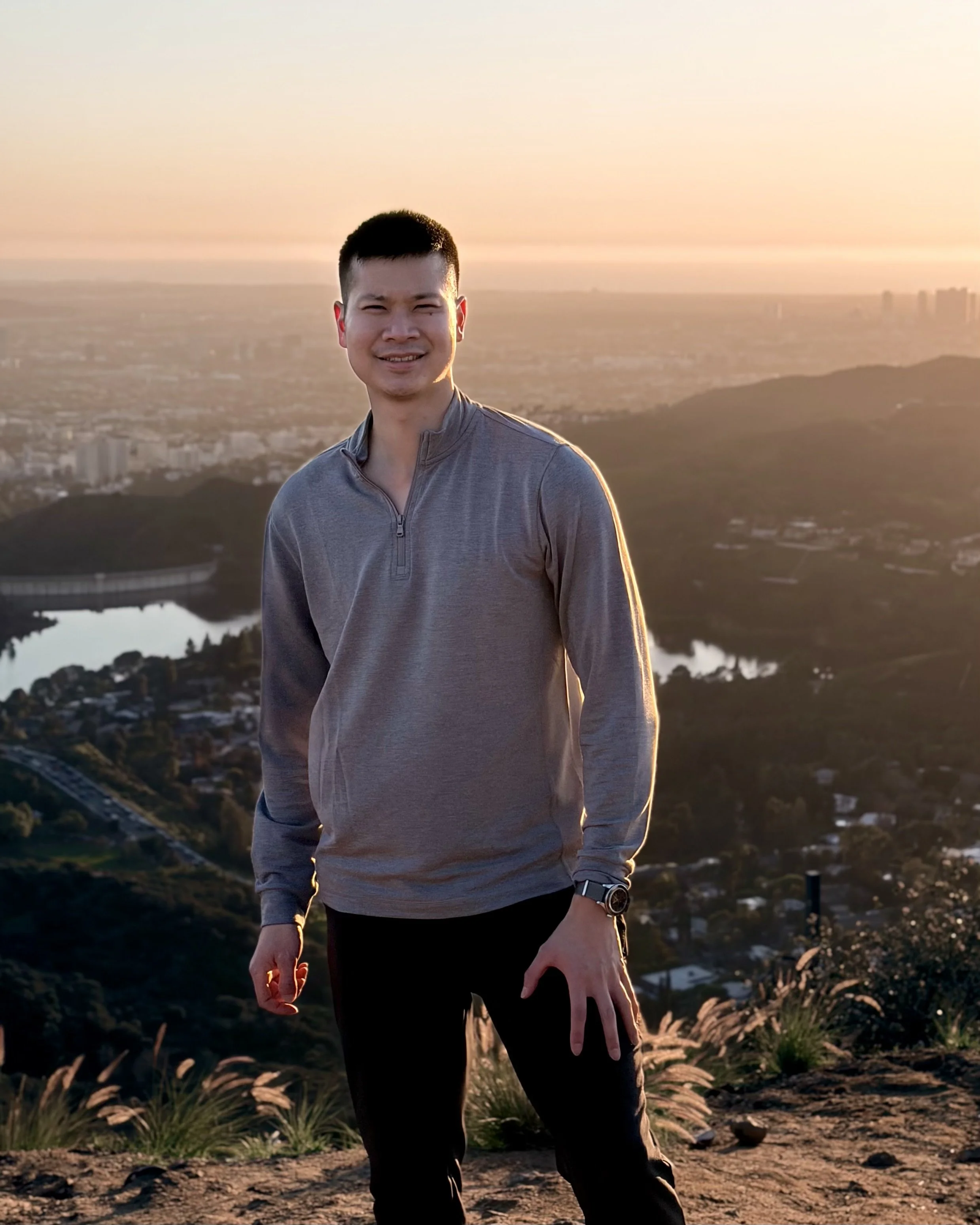 A young man in a gray long-sleeve shirt standing outdoors during sunset, with hills, water, and a cityscape in the background.