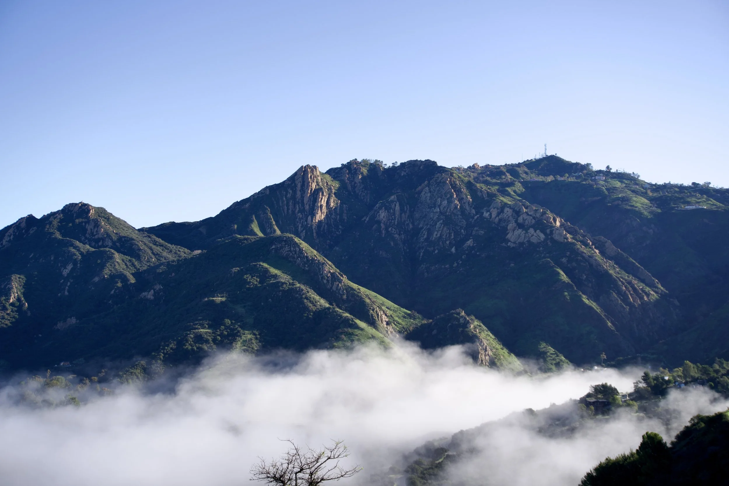 Green mountains with rocky peaks under a clear blue sky, fog covering the lower parts of the mountains.
