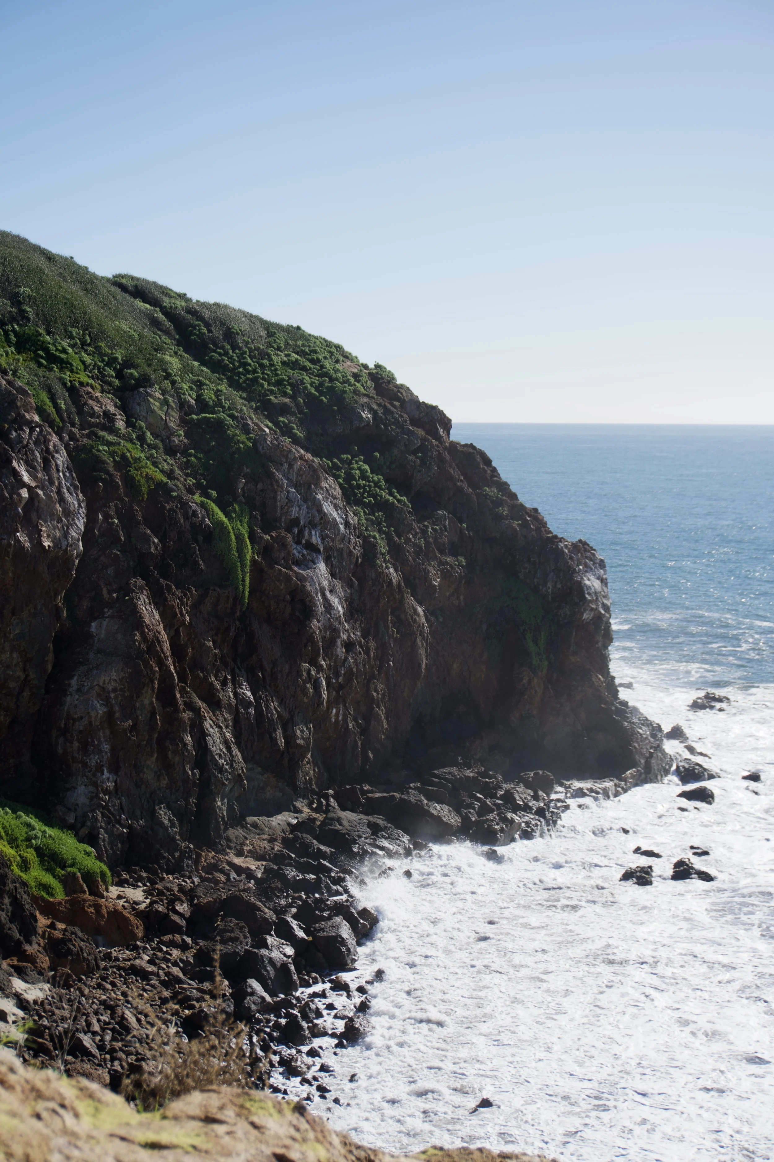 A rocky coastline with green vegetation on cliffs and waves crashing below under a clear blue sky.