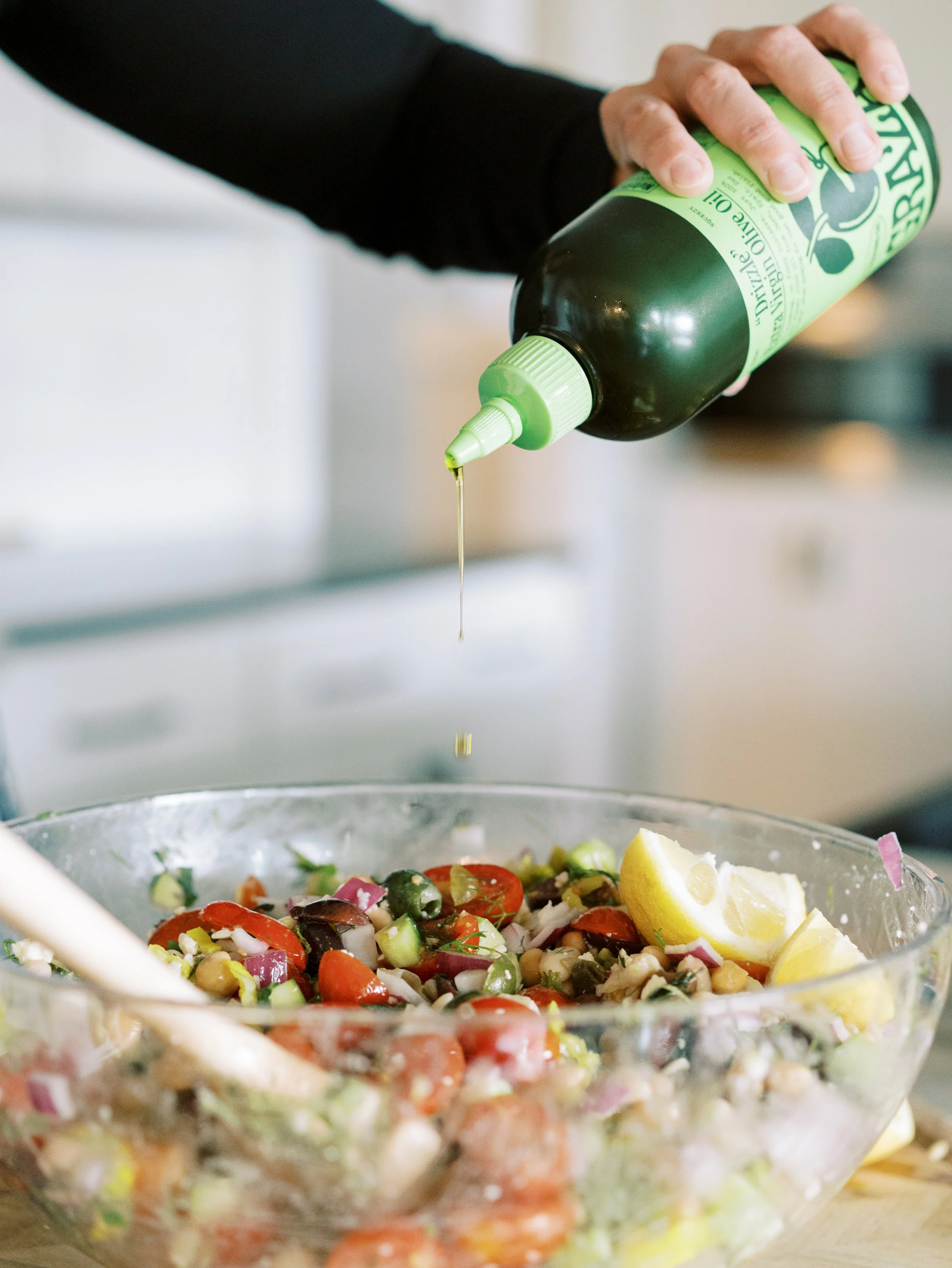 Chef drizzling olive oil over a fresh Mediterranean-style salad with tomatoes, herbs, and lemon.
