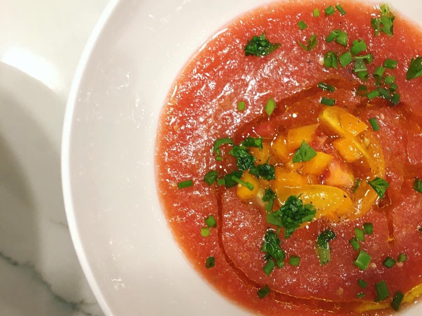 A bowl of tomato soup garnished with chopped green herbs and small chunks of yellow and pink vegetables.
