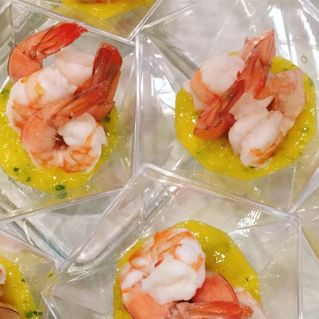 Close-up of shrimp appetizers in clear square cups, topped with yellow sauce and garnished with chopped herbs.