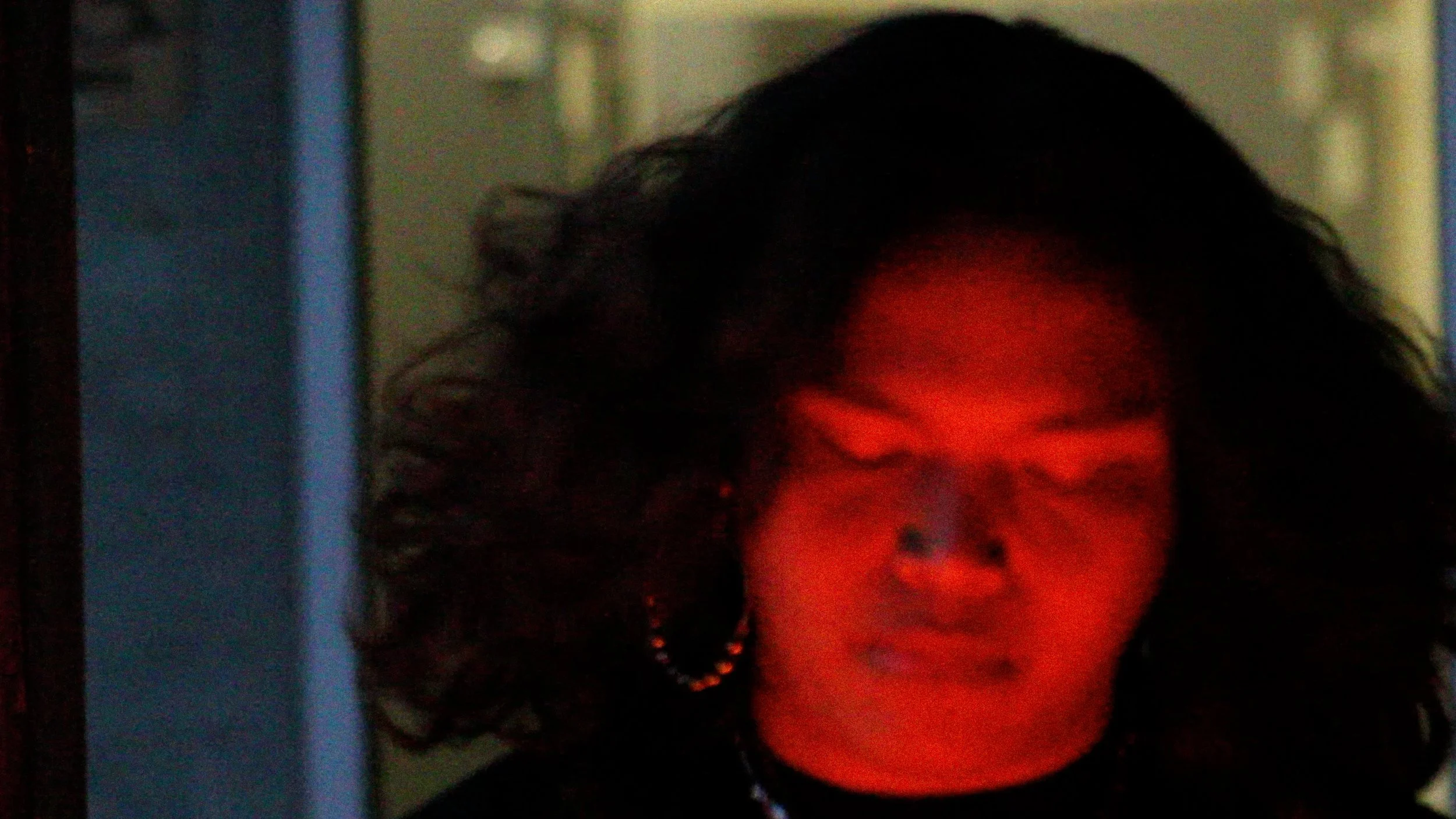 A person with dark, curly hair and a nose piercing, illuminated by red light, in a dark indoor setting.