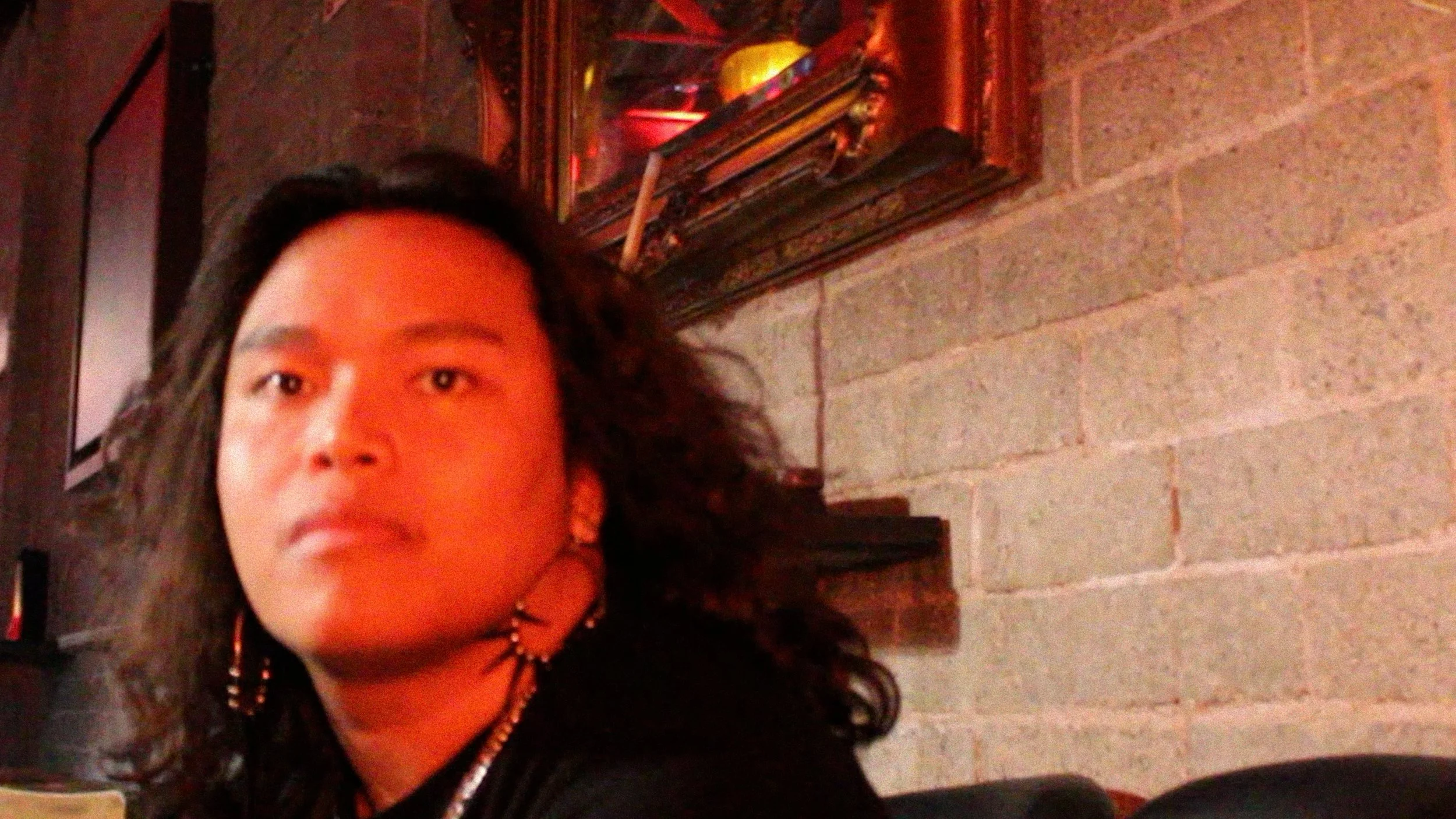 A close-up of a persons face with dark, curly hair, wearing earrings and a necklace, standing against a brick wall with a portrait above their head.
