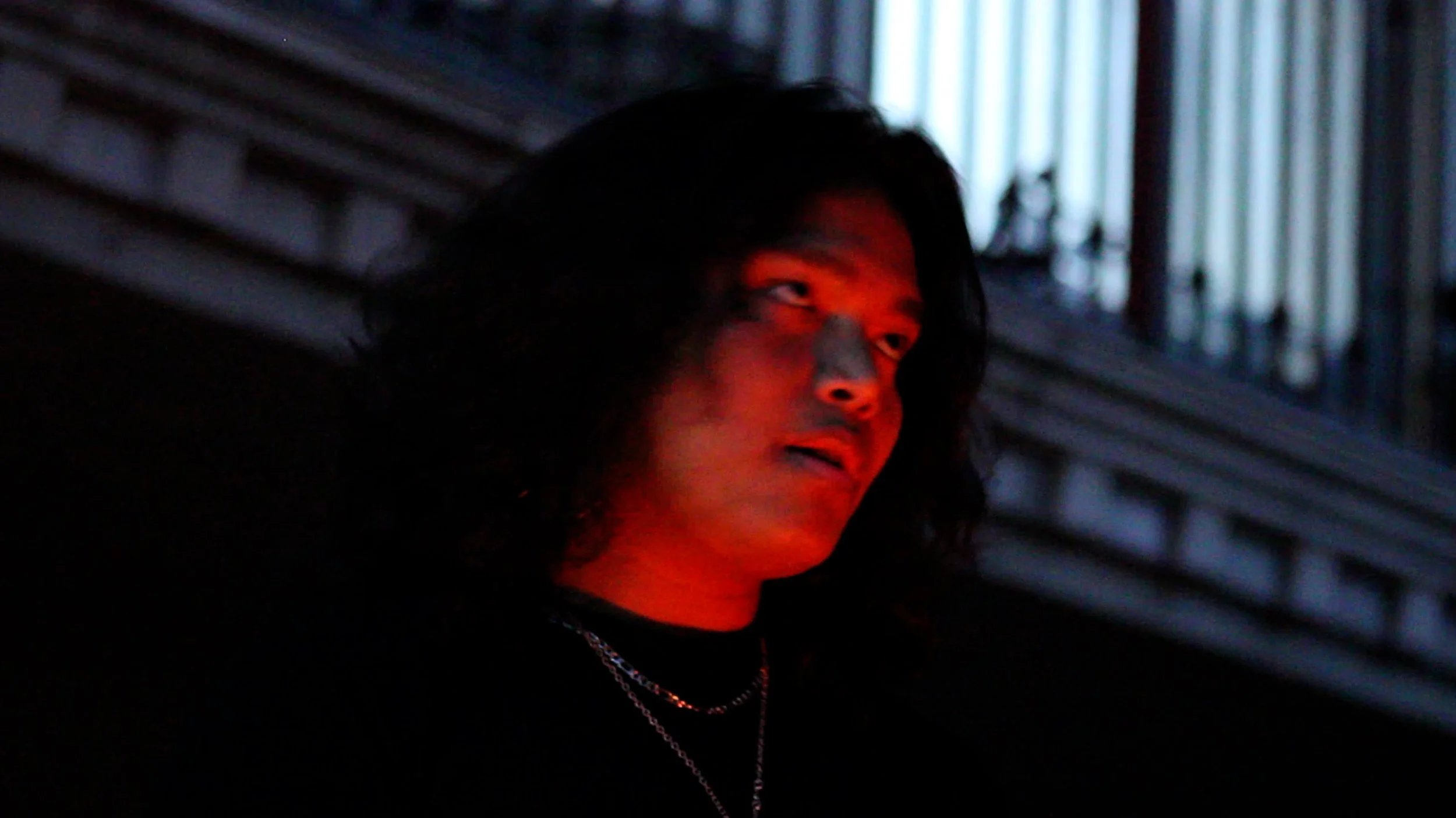 A person with long dark hair looking to the side with a red light illuminating their face in a dark setting.