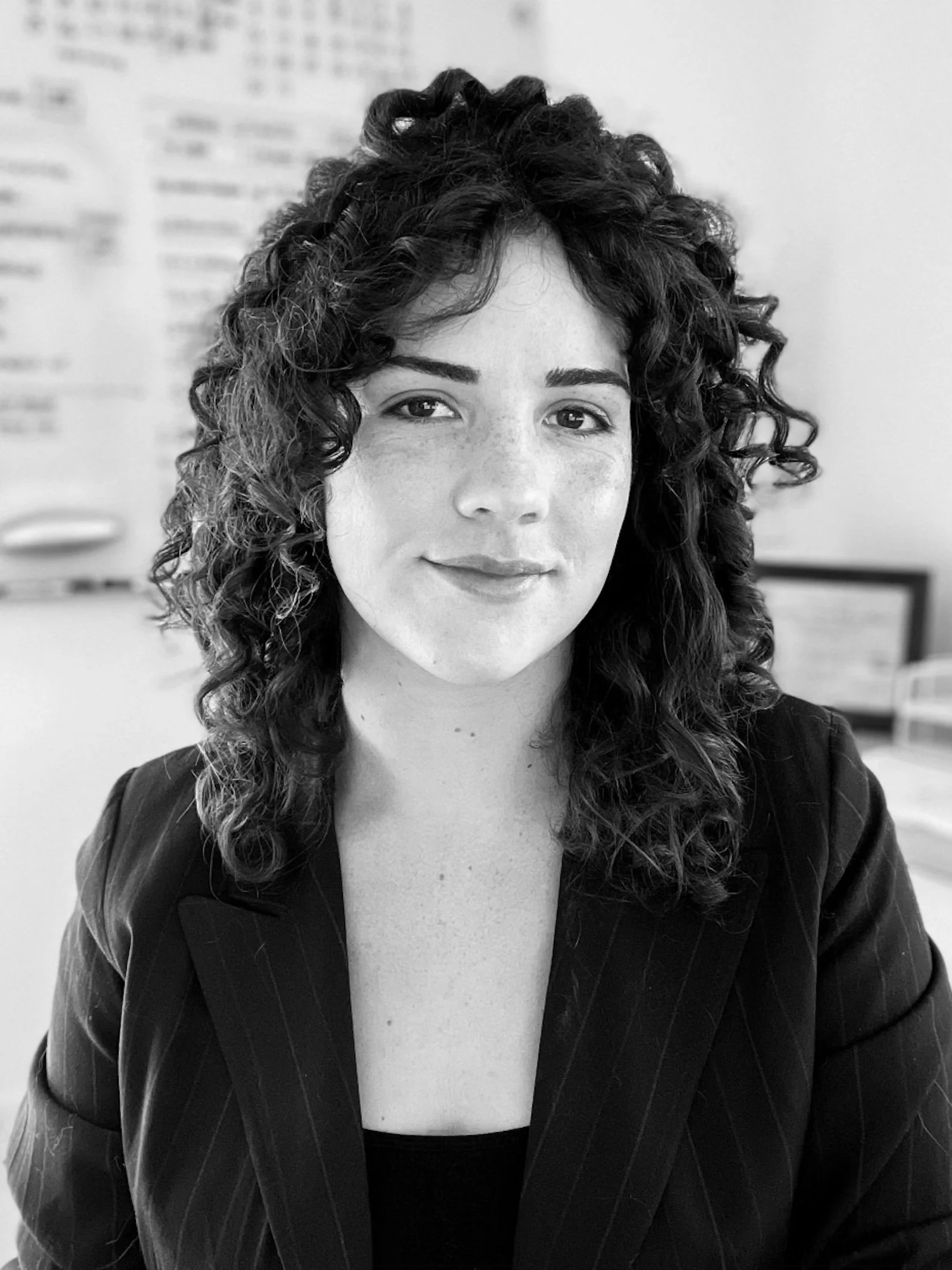 Black and white portrait of a woman with curly hair, wearing a blazer, sitting at a desk in an office environment.