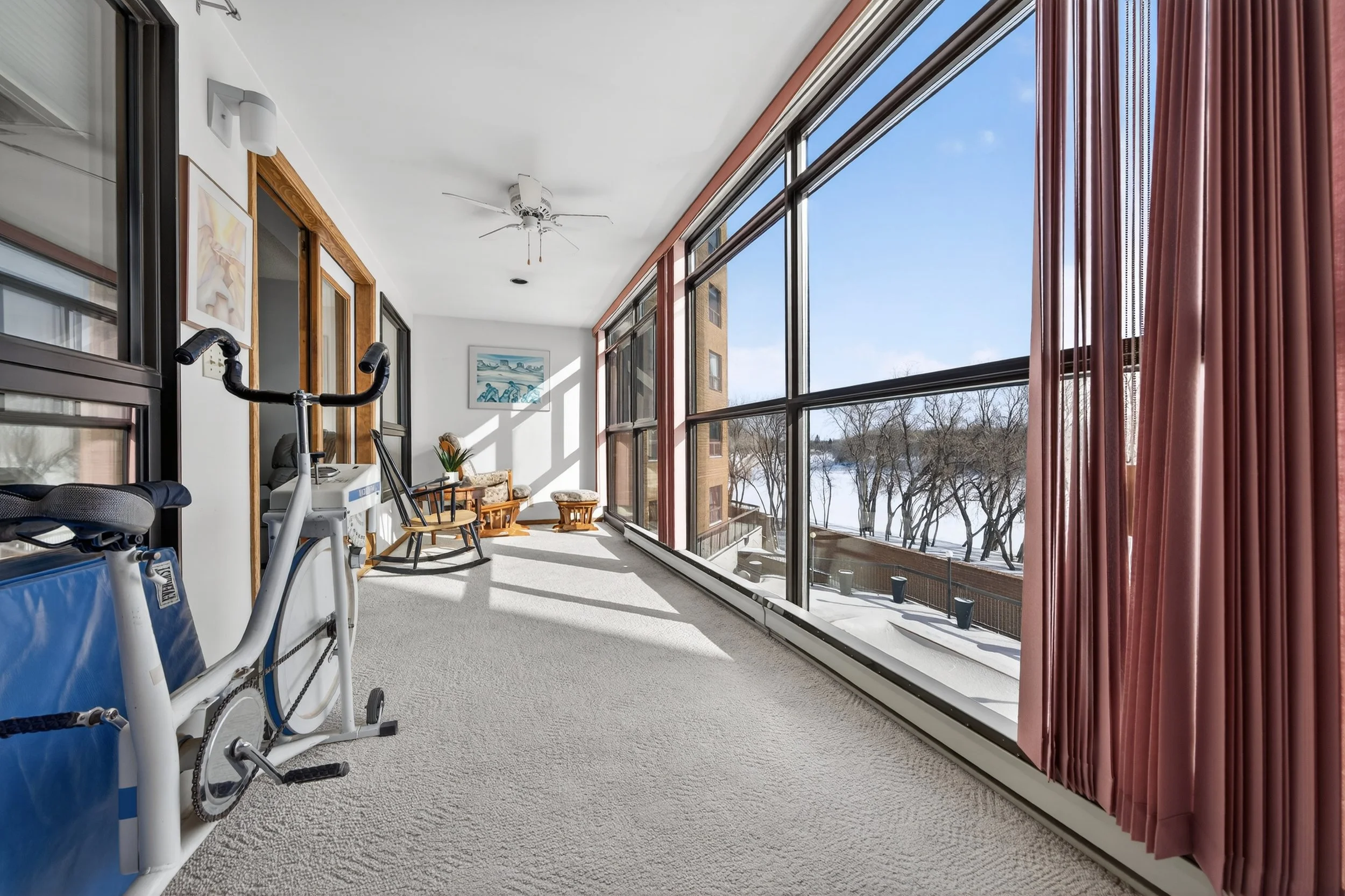Enclosed balcony or sunroom with large windows, exercise bike, wooden rocking chair, small side table with flowers, and a scenic view of snow-covered trees and a lake outside.