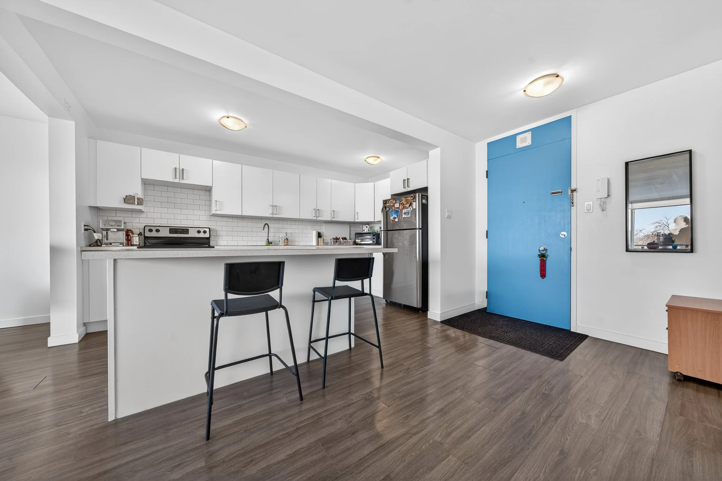 Modern kitchen with white cabinets, a white countertop, a gray refrigerator with magnets, two black barstools, and a blue front door with a small mirror nearby.