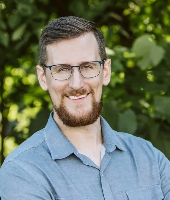 New Page Counselling Clinic Director and Registered Psychotherapist, Nathan Kalk, with glasses and a beard smiling outdoors with green foliage in the background.