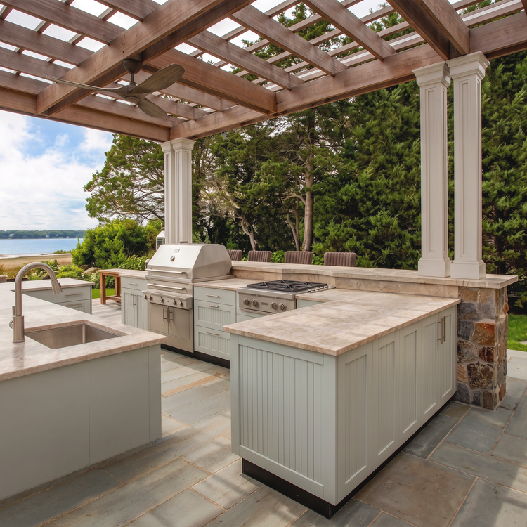 A coastal outdoor kitchen in pale cabinetry with a built-in BBQ, stone bench tops and a sea view beyond