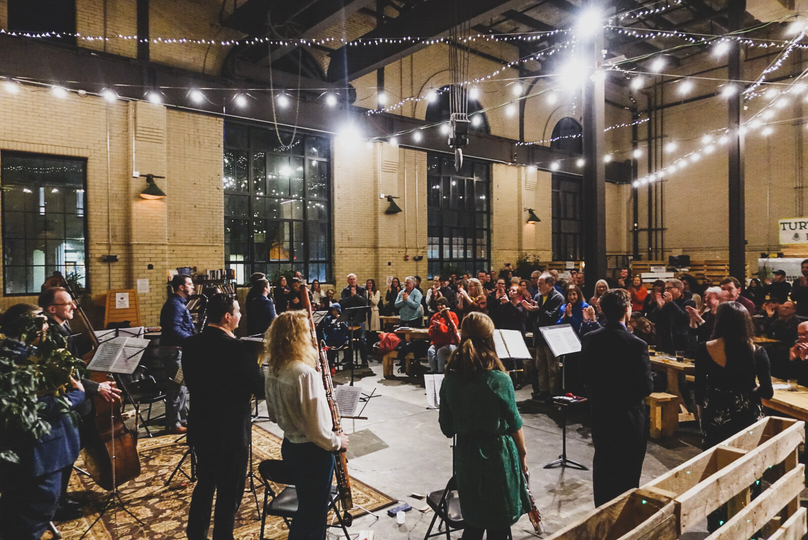 A musical performance with a group playing instruments in front of an audience inside a spacious industrial-style venue with high ceilings, large windows, and string lights.