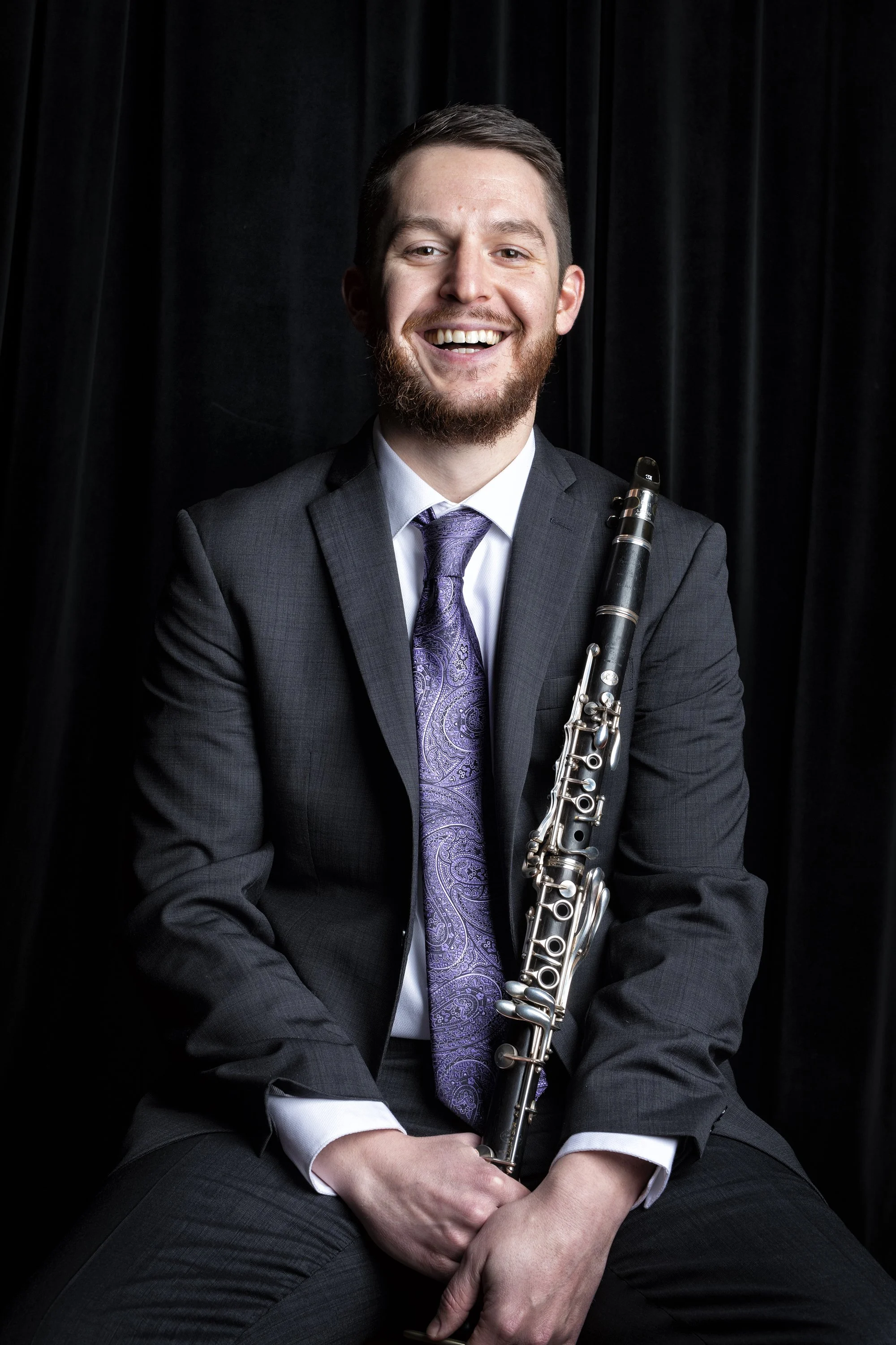A young man in a suit and purple tie, holding a clarinet, smiling, against a black curtain background.