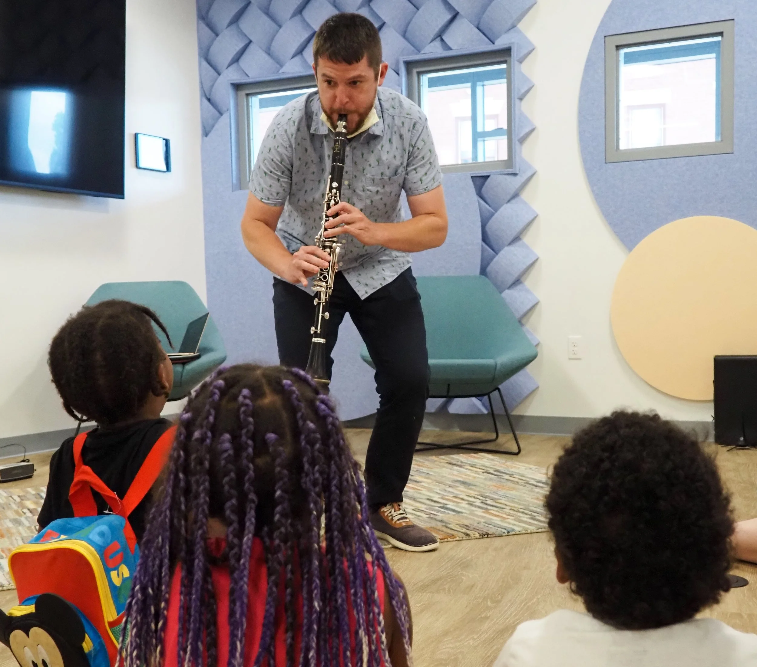 A man is playing a clarinet in a music classroom with children seated on the floor watching him.