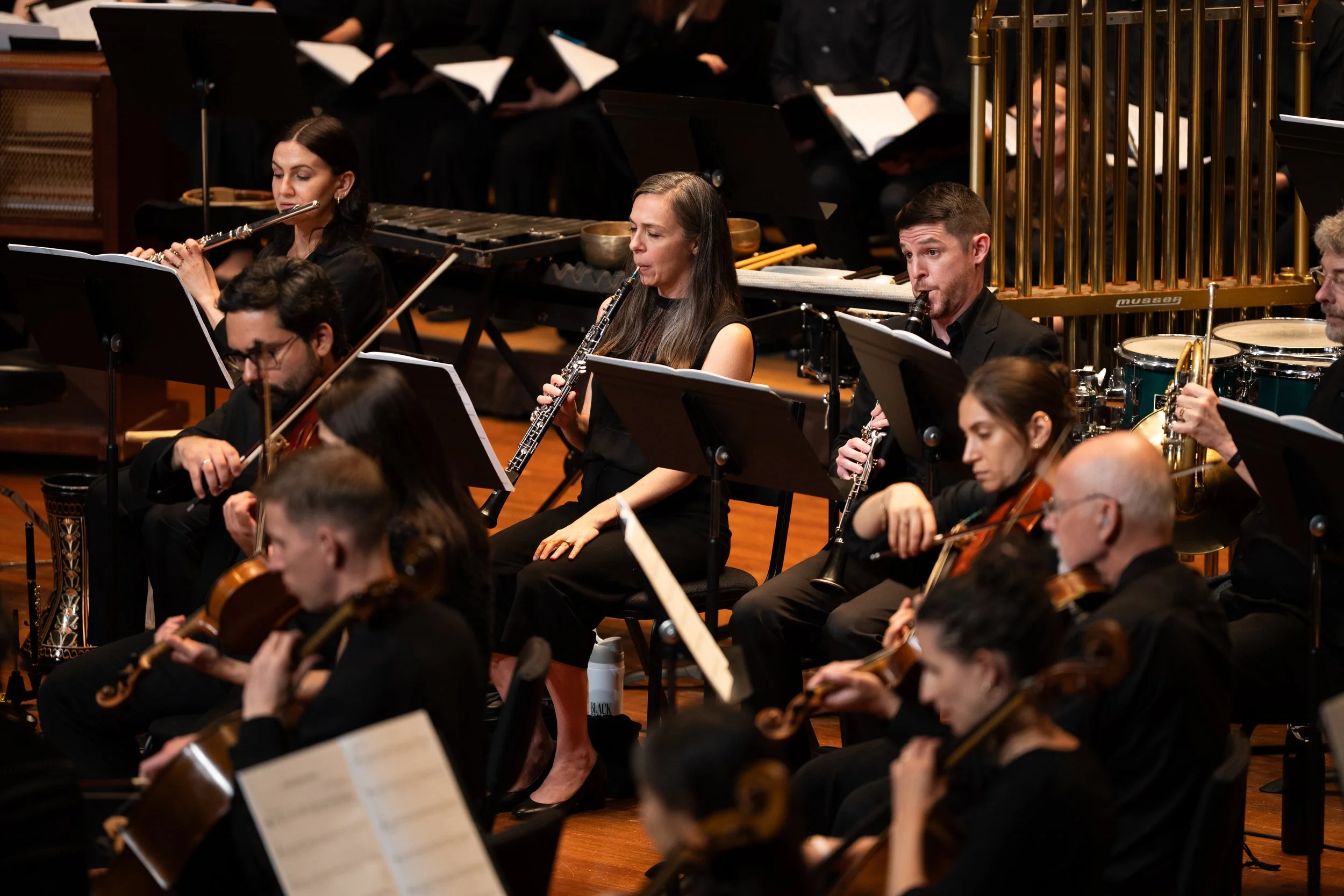Orchestra performing on stage in a concert hall, with musicians playing various instruments including violins, cellos, and woodwinds.