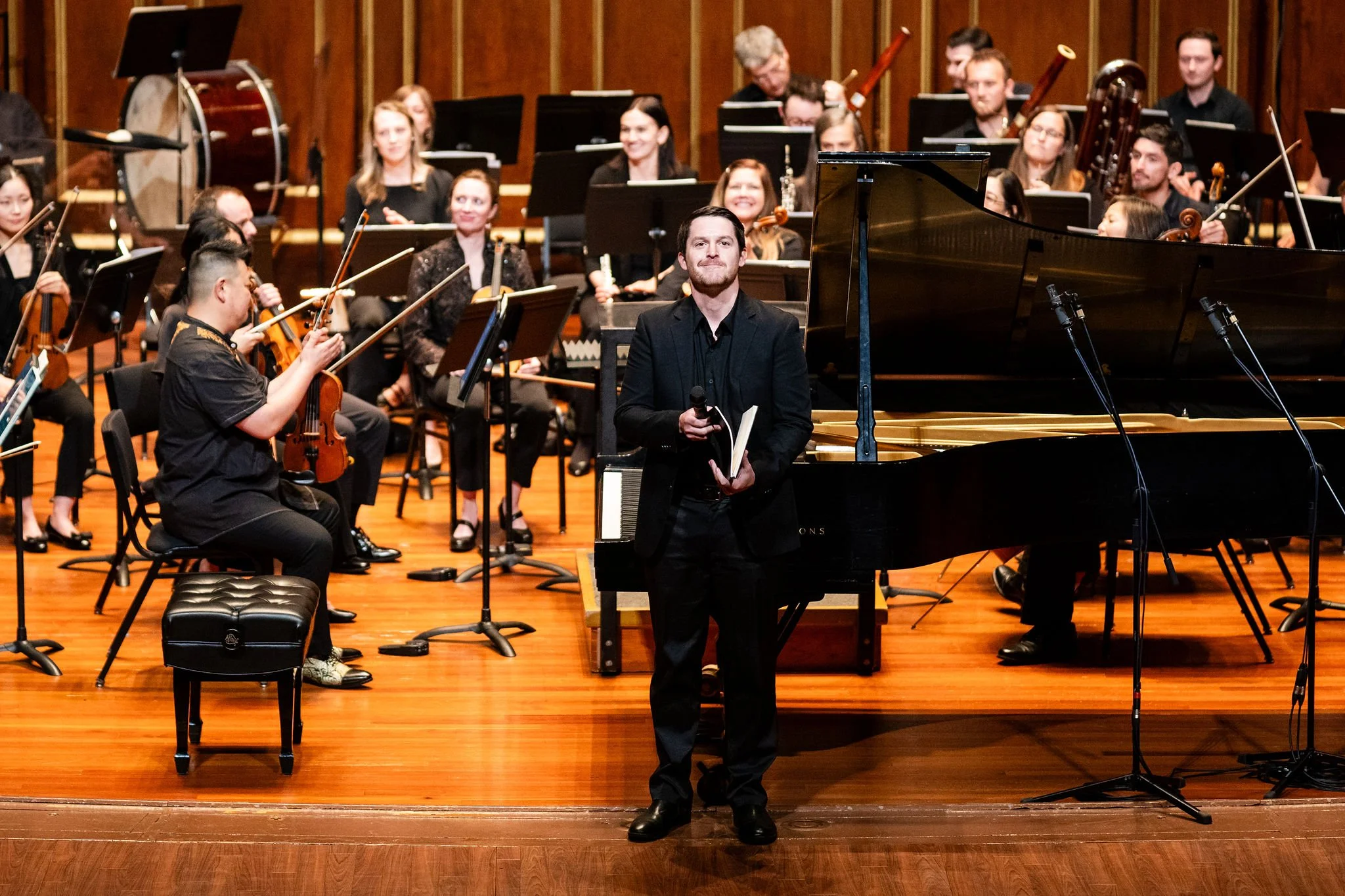 Orchestra conductor stands by grand piano on stage, holding a microphone and a notebook, with a large orchestra seated and playing string, wind, and percussion instruments in the background.