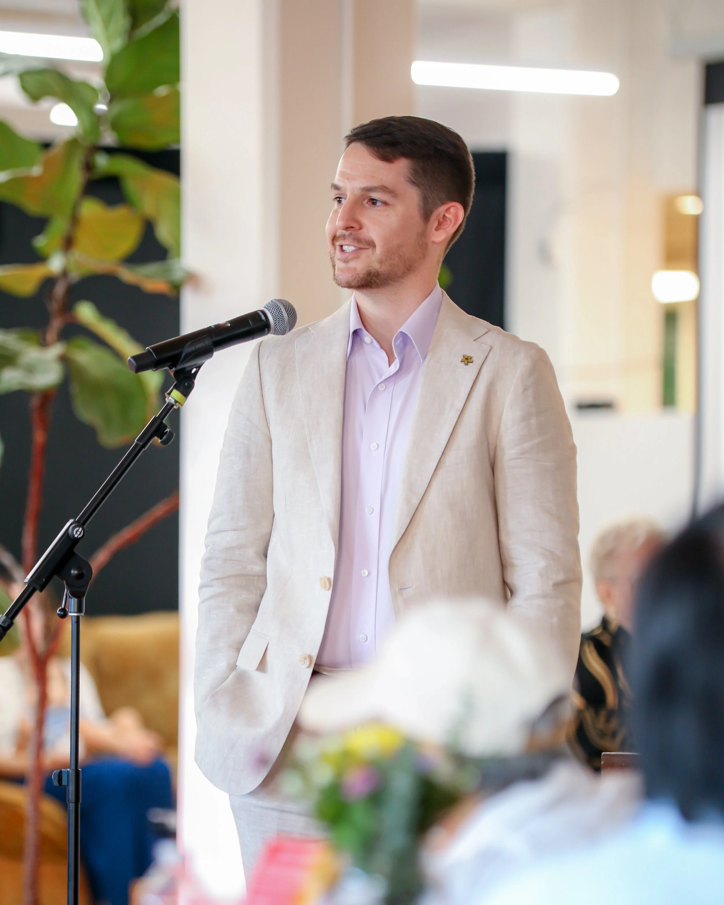 A man in a cream-colored suit speaking at a microphone during an event. There are blurred flowers and people in the background.