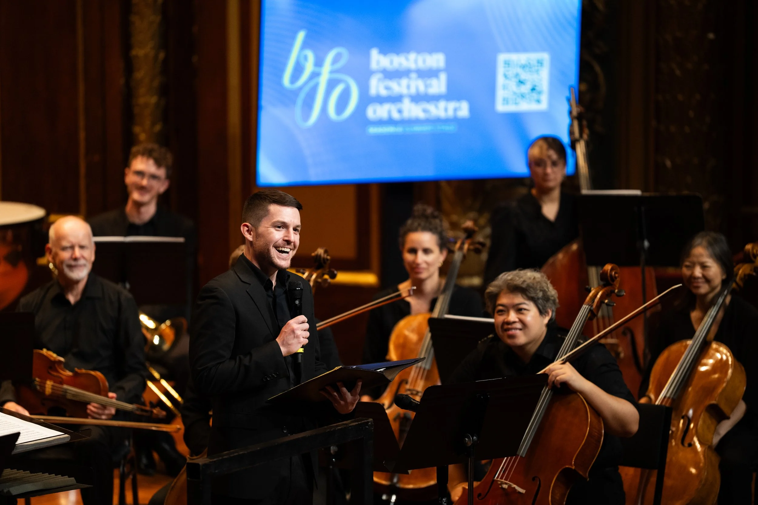 A young man in a black suit holding a microphone and smiling, standing in front of an orchestra with musicians playing string instruments, at a concert hall with a large screen displaying 'Boston Festival Orchestra' in the background.