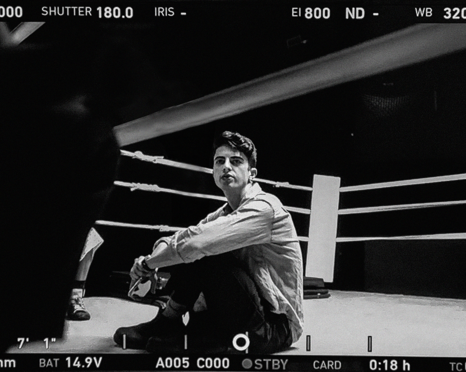 Black and white photo of a person sitting on the floor of a boxing ring, with ringside ropes behind them, and camera interface elements overlaid.