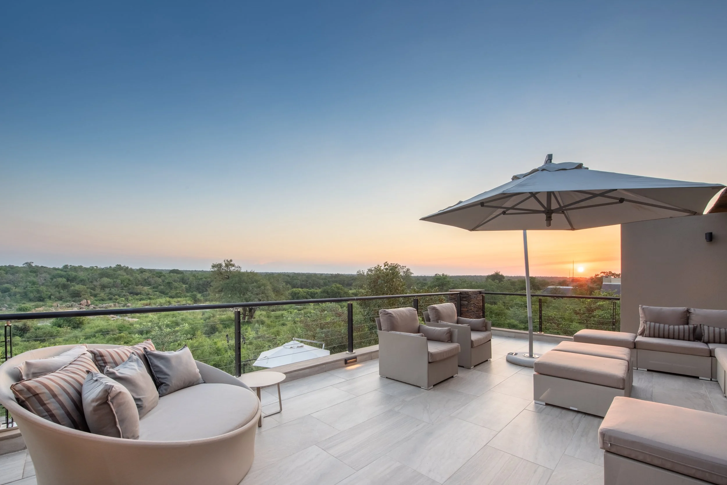 Outdoor patio furniture with cushioned chairs and sofas, a white umbrella, and a view of a green landscape at sunset.