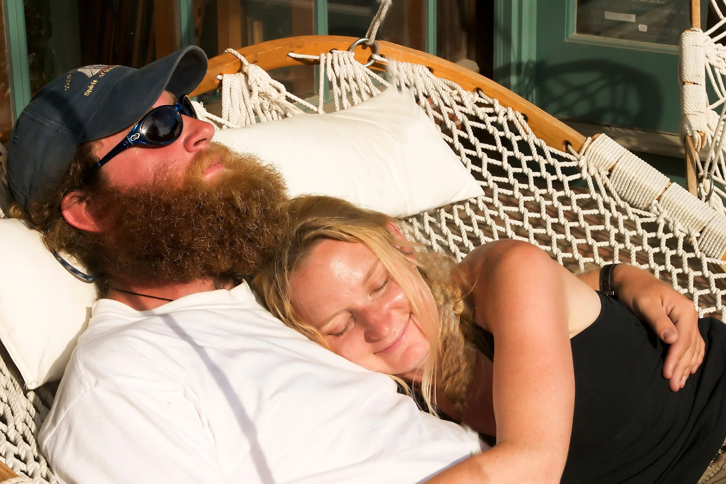 Man with beard, sunglasses and hat, and a blond woman in a black tank top relax in a hammock.