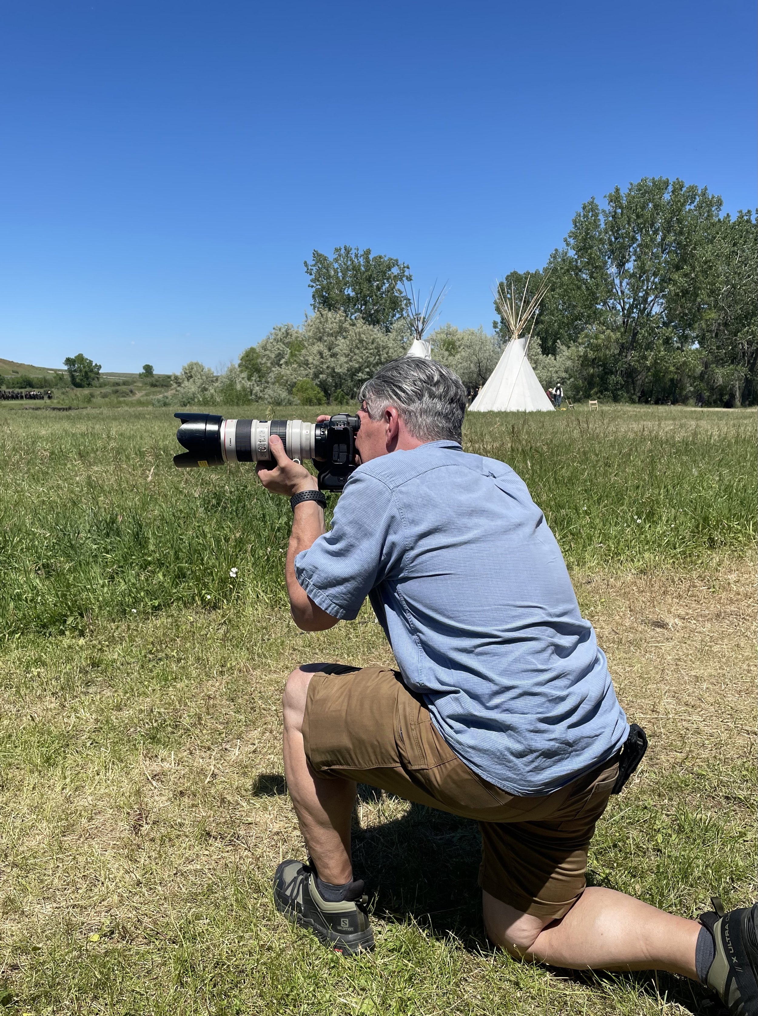 Jon kneeling on grass taking a photograph with a professional camera in a field with green trees and white tipi tents in the background under a clear blue sky.