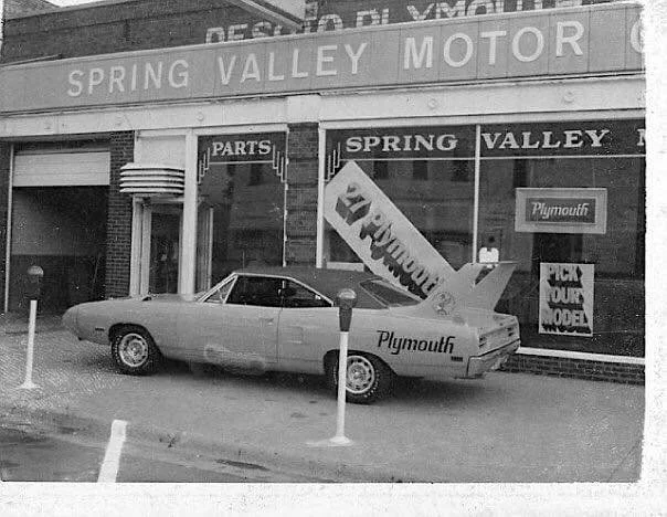 A vintage Plymouth car with a large rear fin parked in front of a brick storefront. The store has signs for Spring Valley Motor, parts, and Plymouth, with banners and posters in the windows advertising cars and parts.