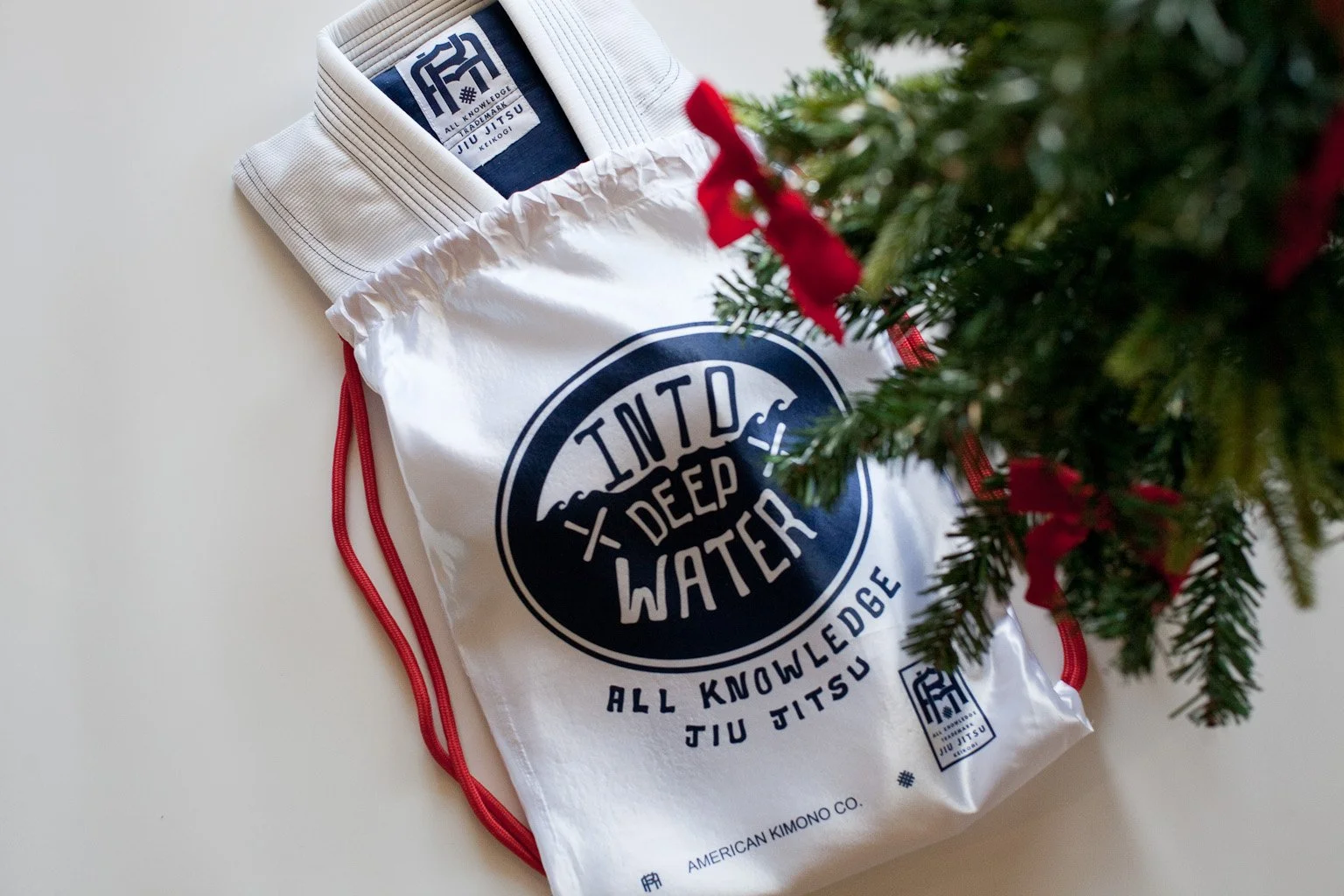 A white drawstring bag with a black logo reading 'INTO DEEP WATER' and 'ALL KNOWLEDGE JIU JITSU' seen next to a partially visible Christmas tree with red ribbons.