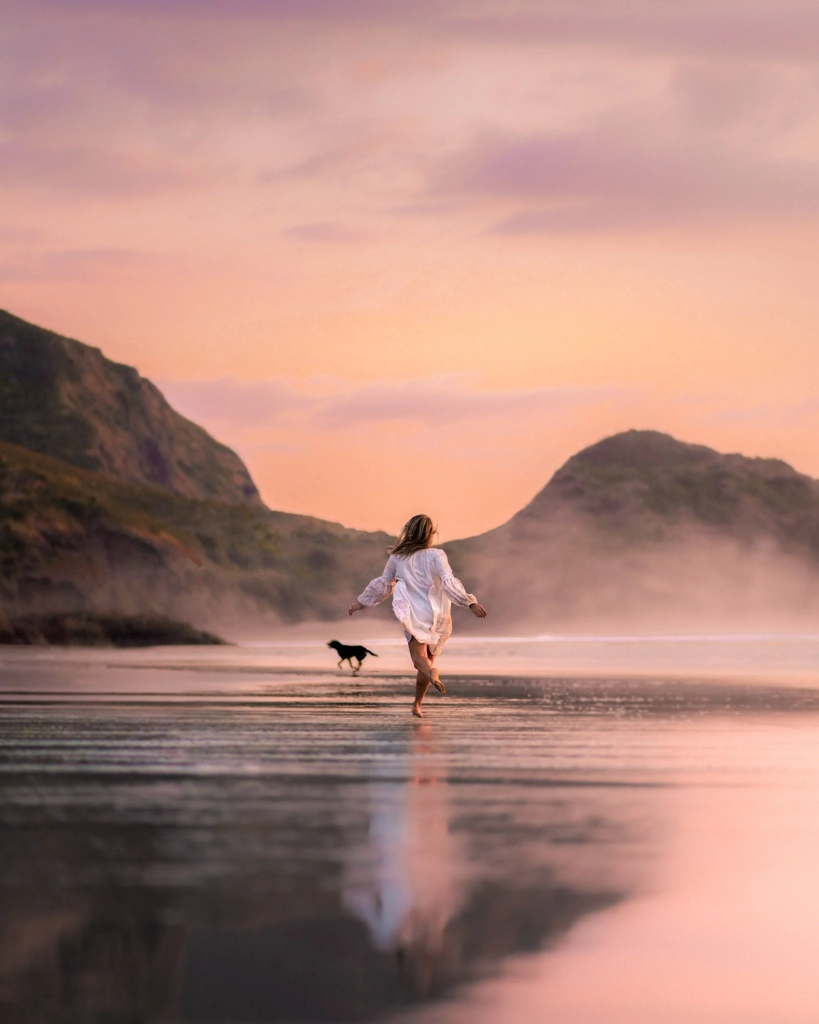 A woman in a white dress running on a beach with a dog, during sunset with hills in the background and a pink sky.