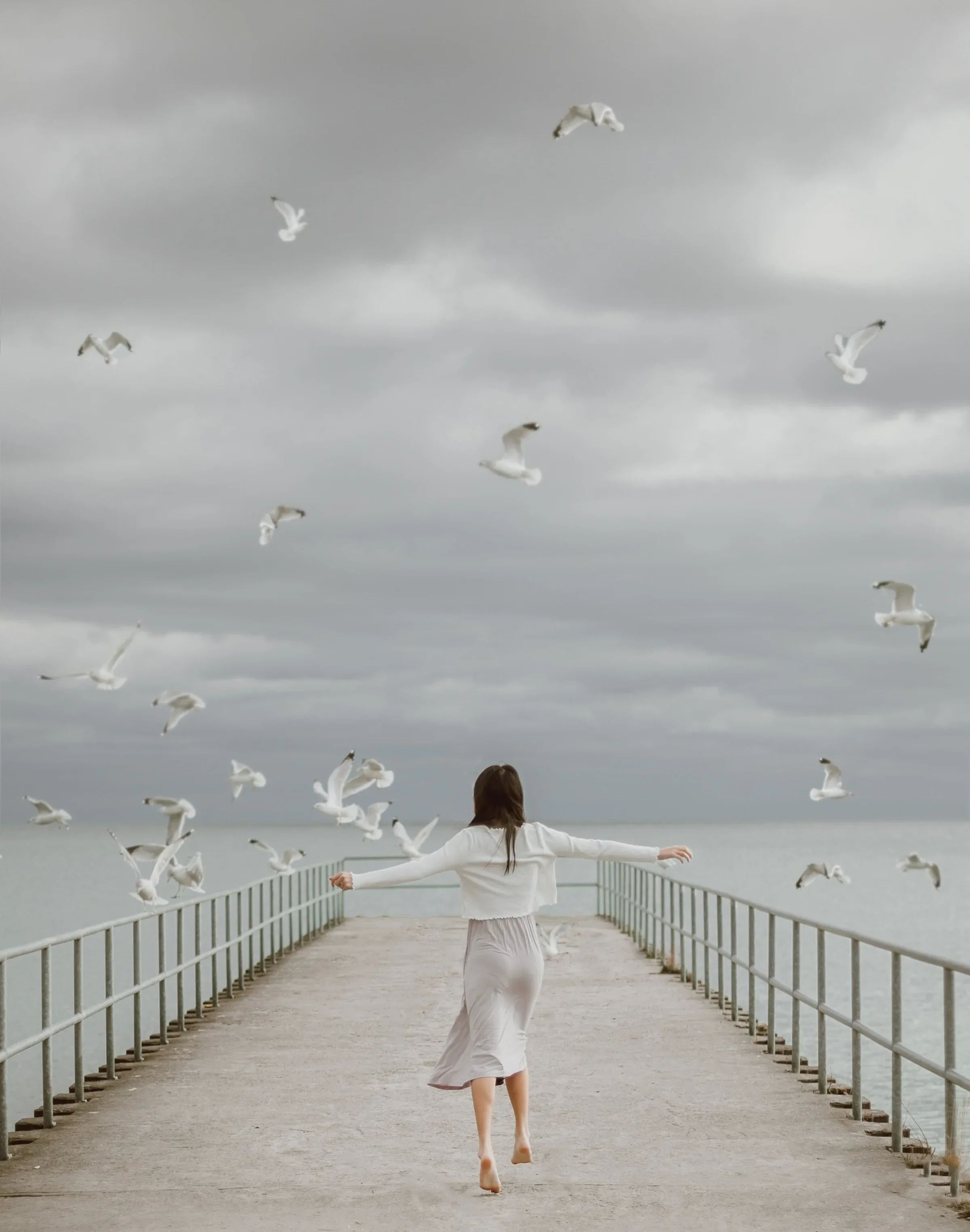 A woman in a white dress and sweater running on a pier over water, surrounded by flying seagulls on a cloudy day.